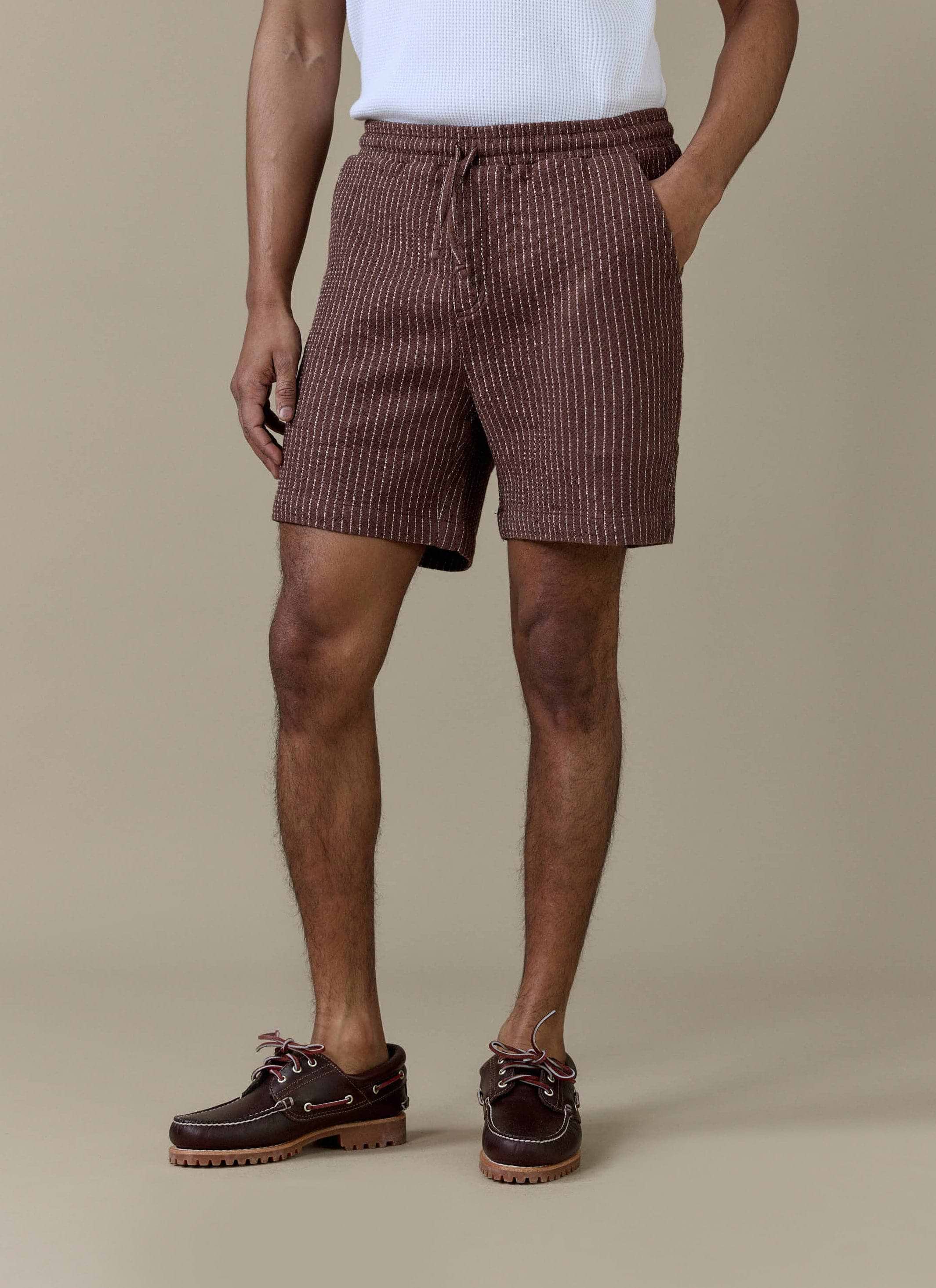 A model wearing the brown pinstripe Sorrento linen shorts from Percival, featuring an elasticated drawstring waistband.