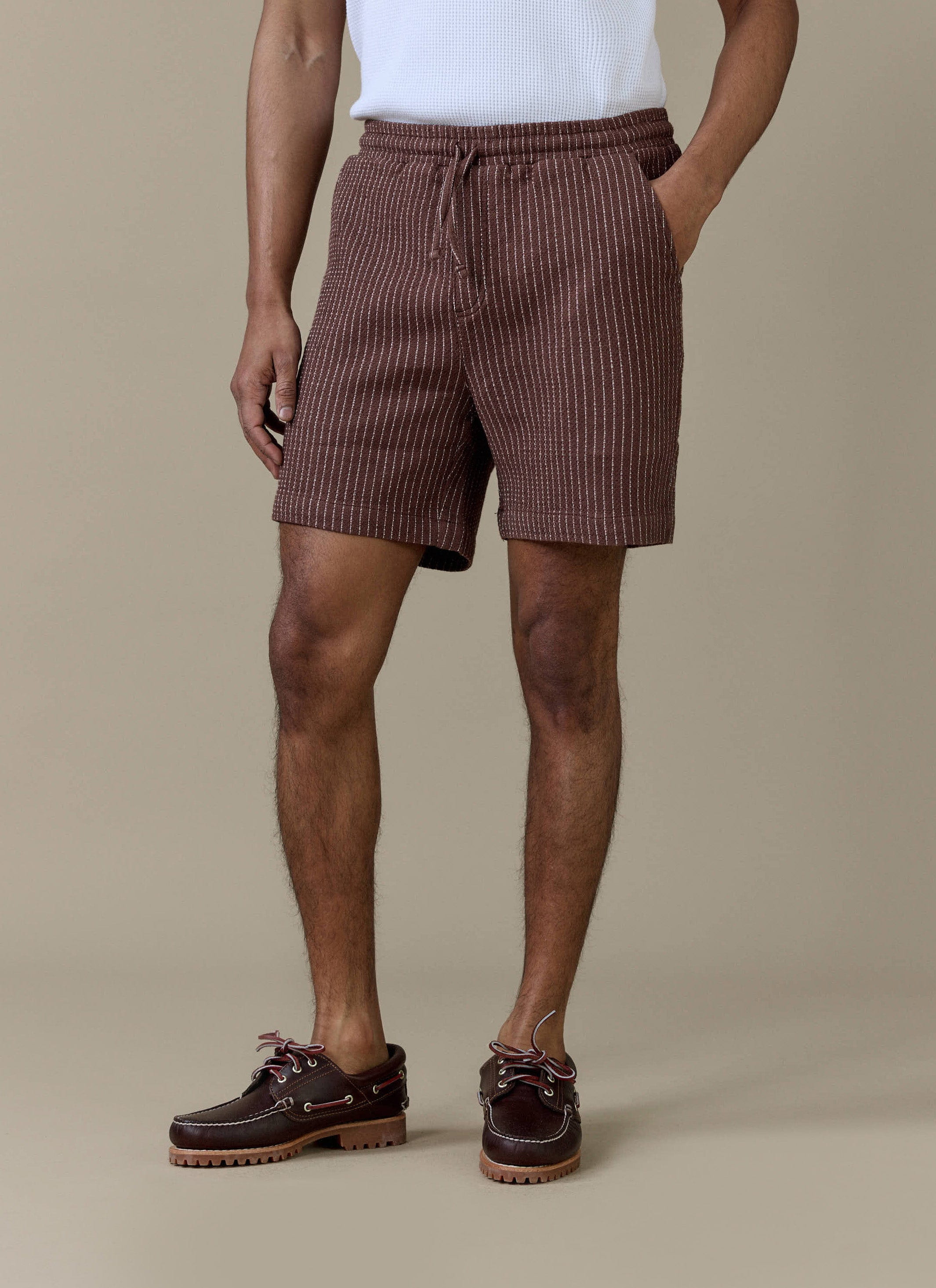 A model wearing the brown pinstripe Sorrento linen shorts from Percival, featuring an elasticated drawstring waistband.