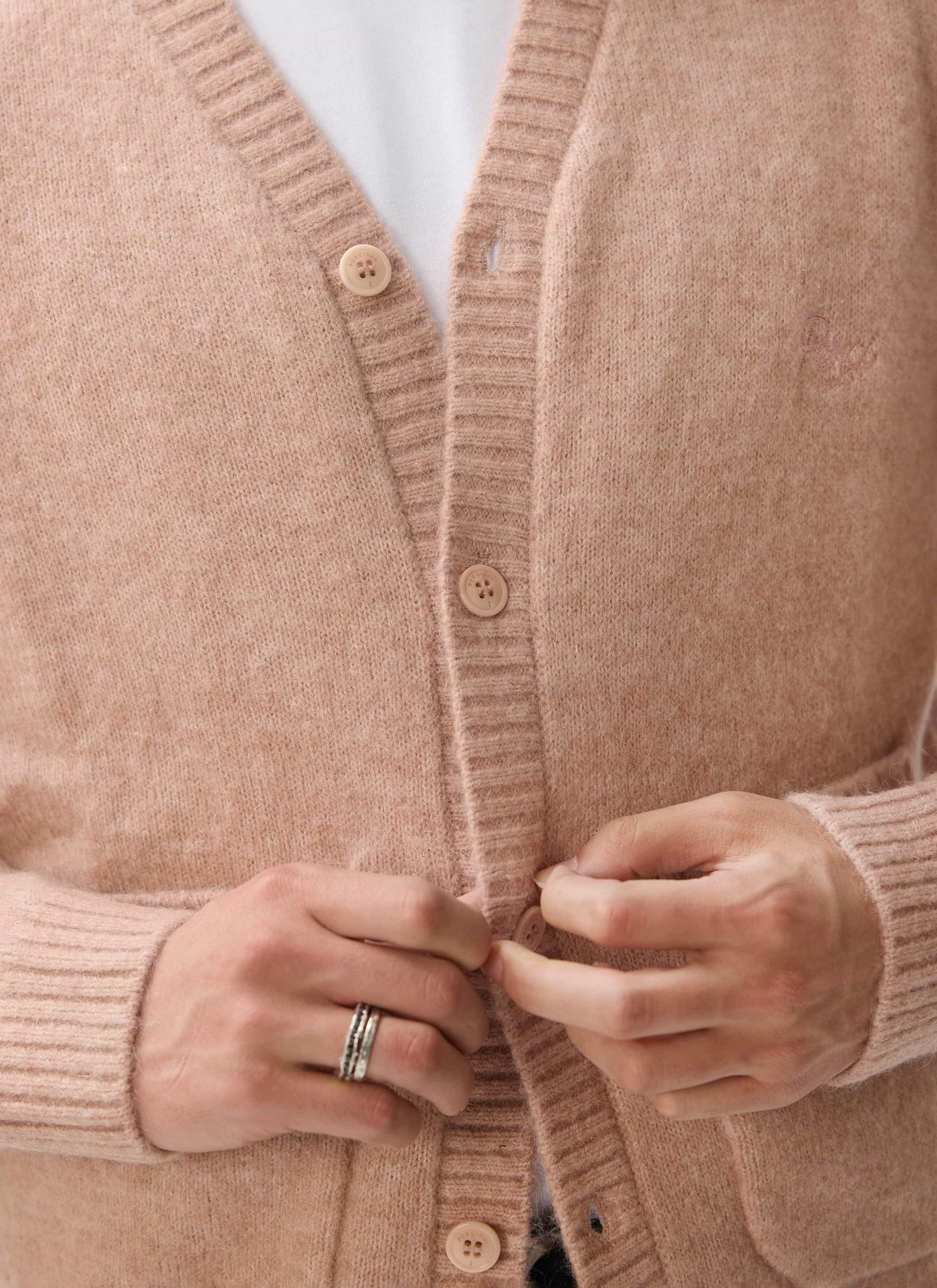A close-up of a model fastening the buttons of the pink mohair White Glove Cardigan by Percival, showing tonal embroidery.