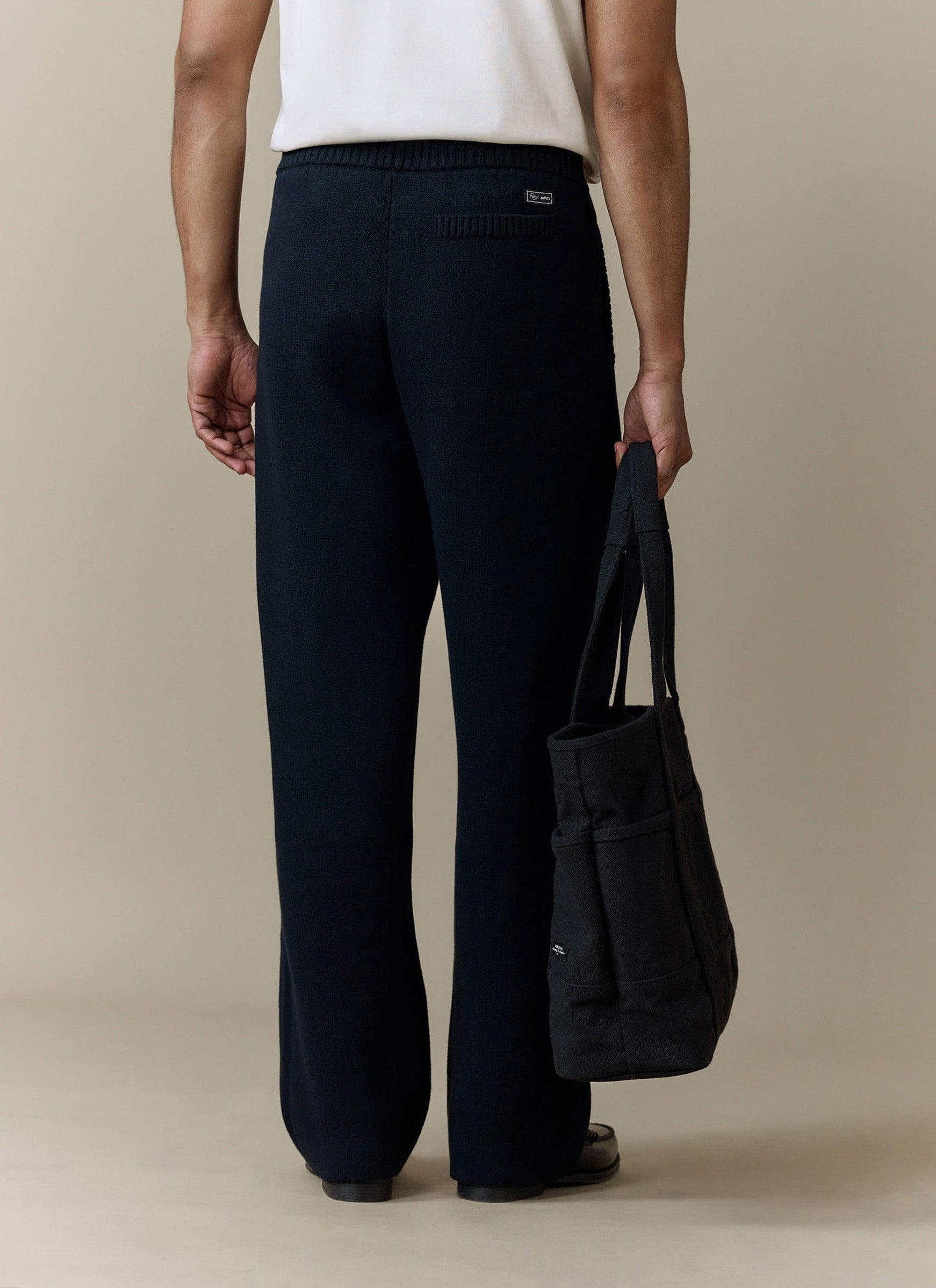 Back view of a model wearing the Percival Chronicle Knitted Straight Leg Trackpant in black, carrying a black tote bag.
