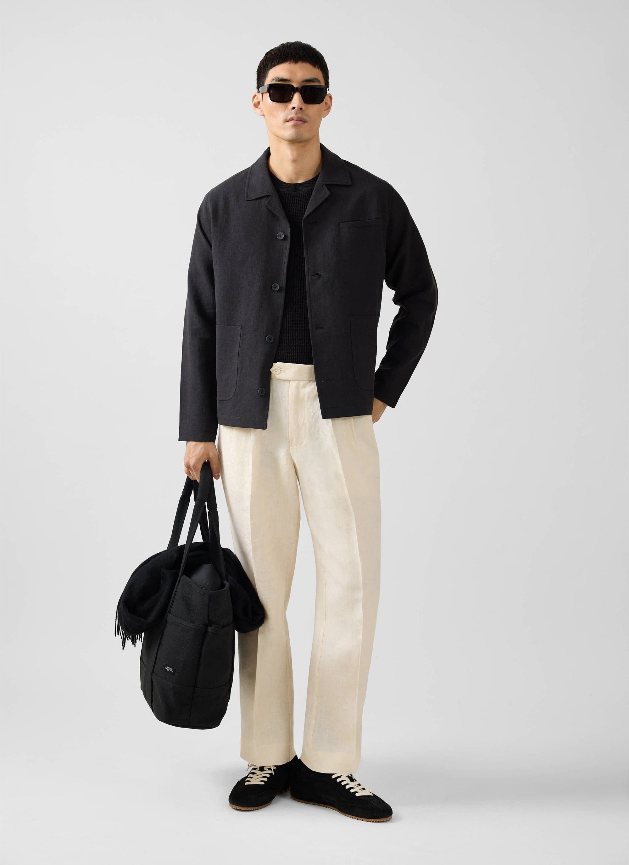 A man models the Percival black linen overshirt, styled with cream trousers, black trainers and sunglasses.