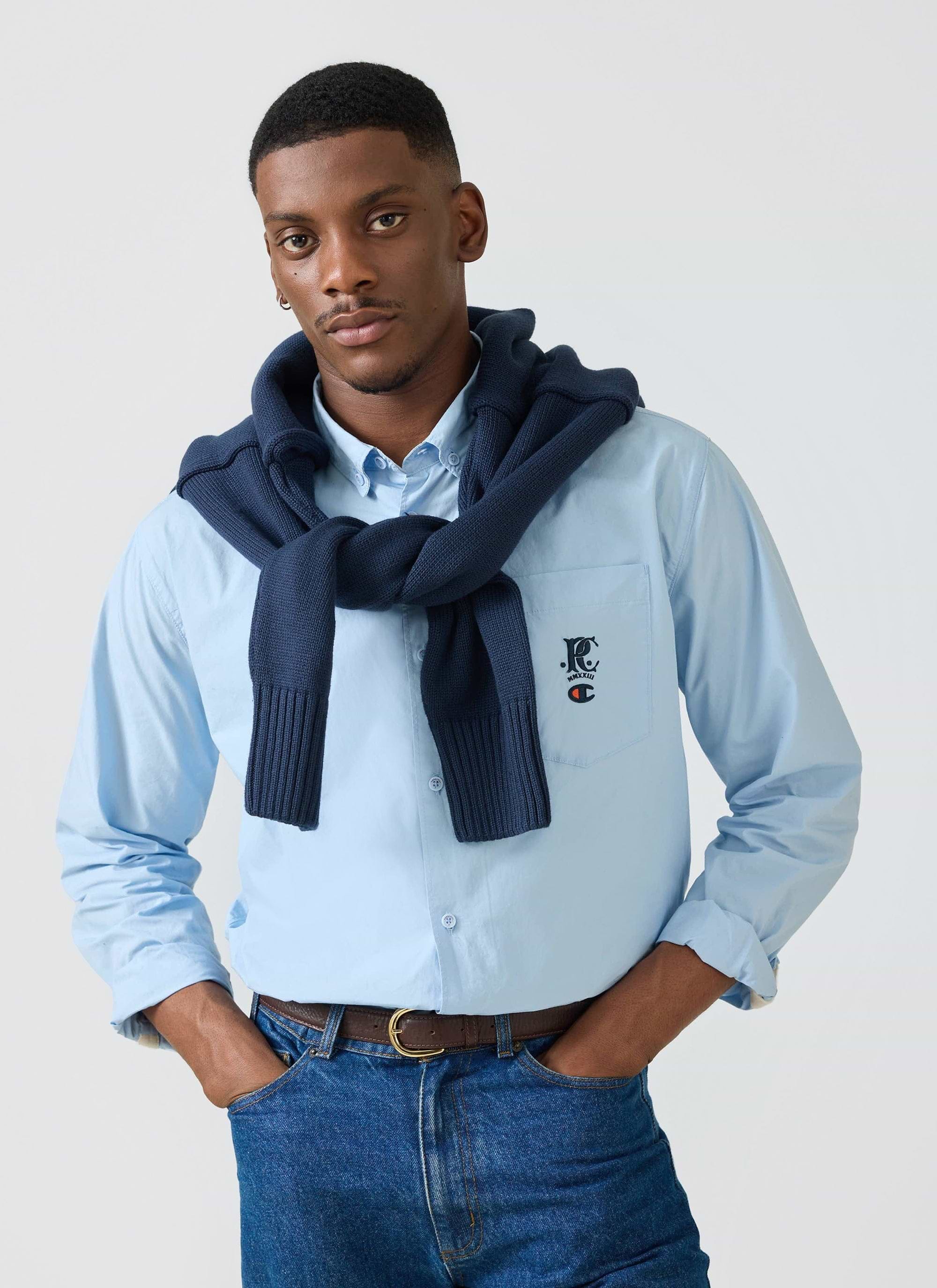 A man models the Champion x Percival Relaxed Oxford Shirt in light blue, with a navy jumper draped over his shoulders.