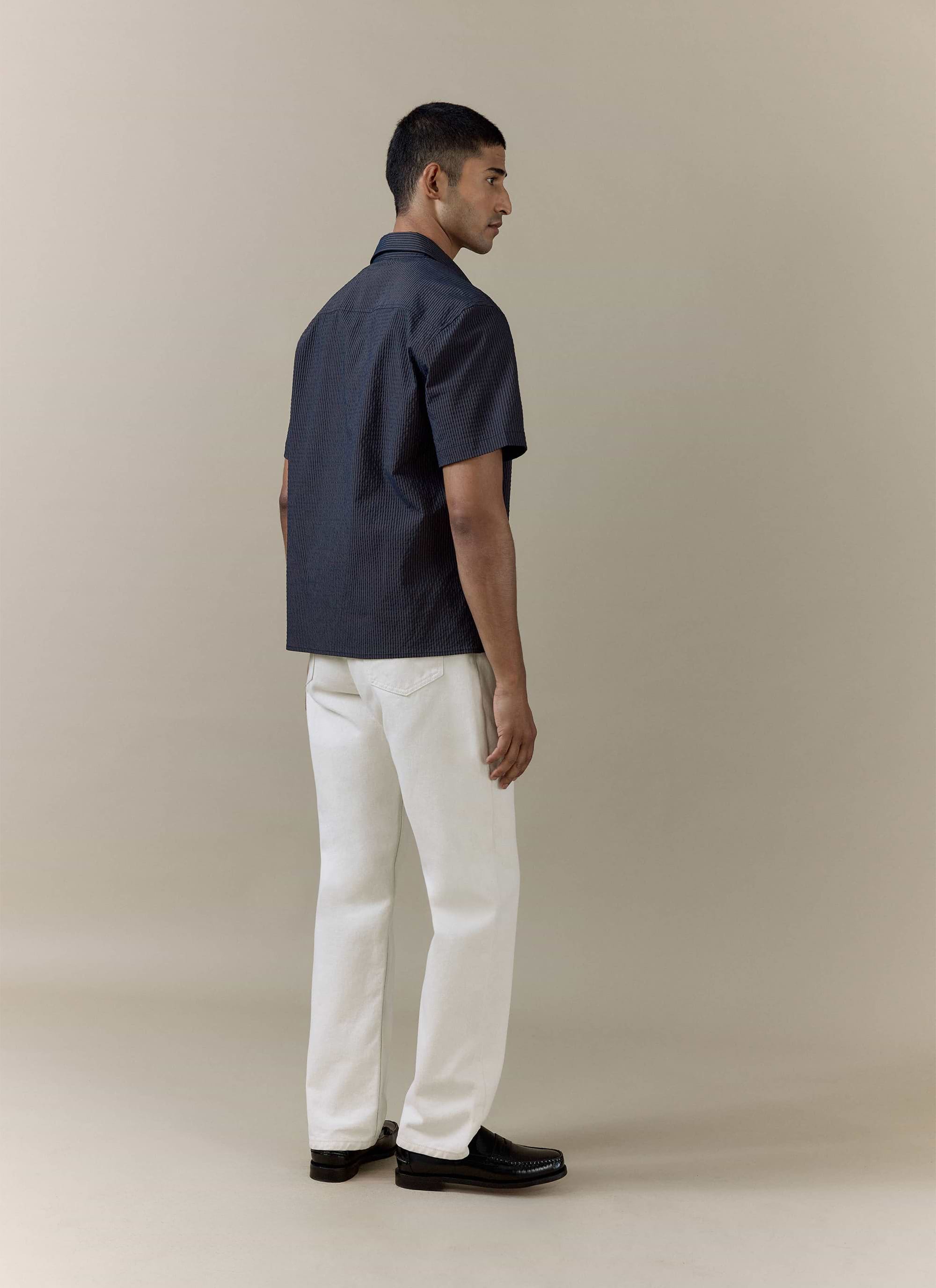 Percival Rowan Seersucker Boxy Shirt in navy stripe, shown from the back with white trousers and black loafers.