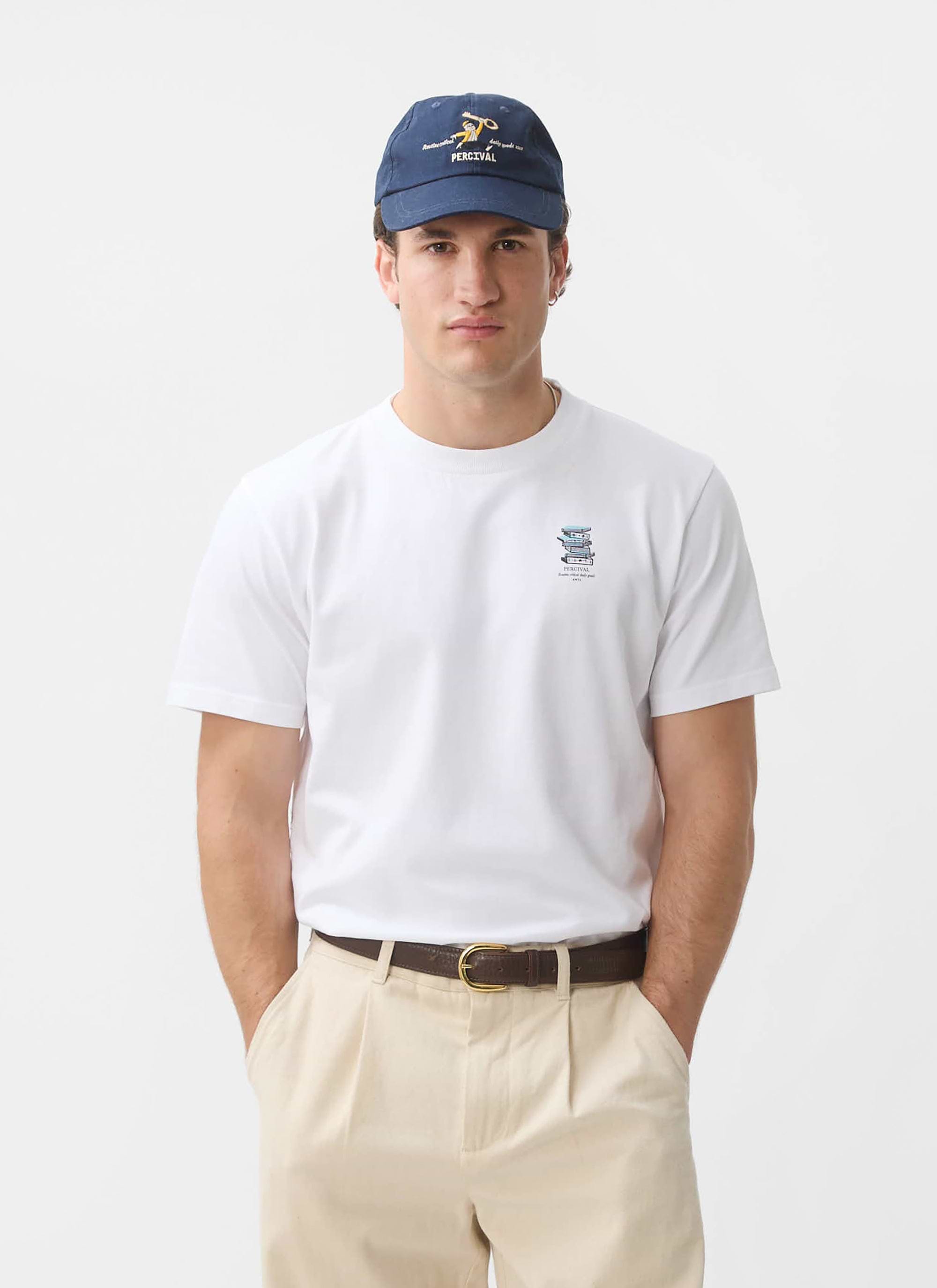 A model wears the white Duke T Shirt from Percival, featuring a book graphic, styled with beige trousers and a blue cap.