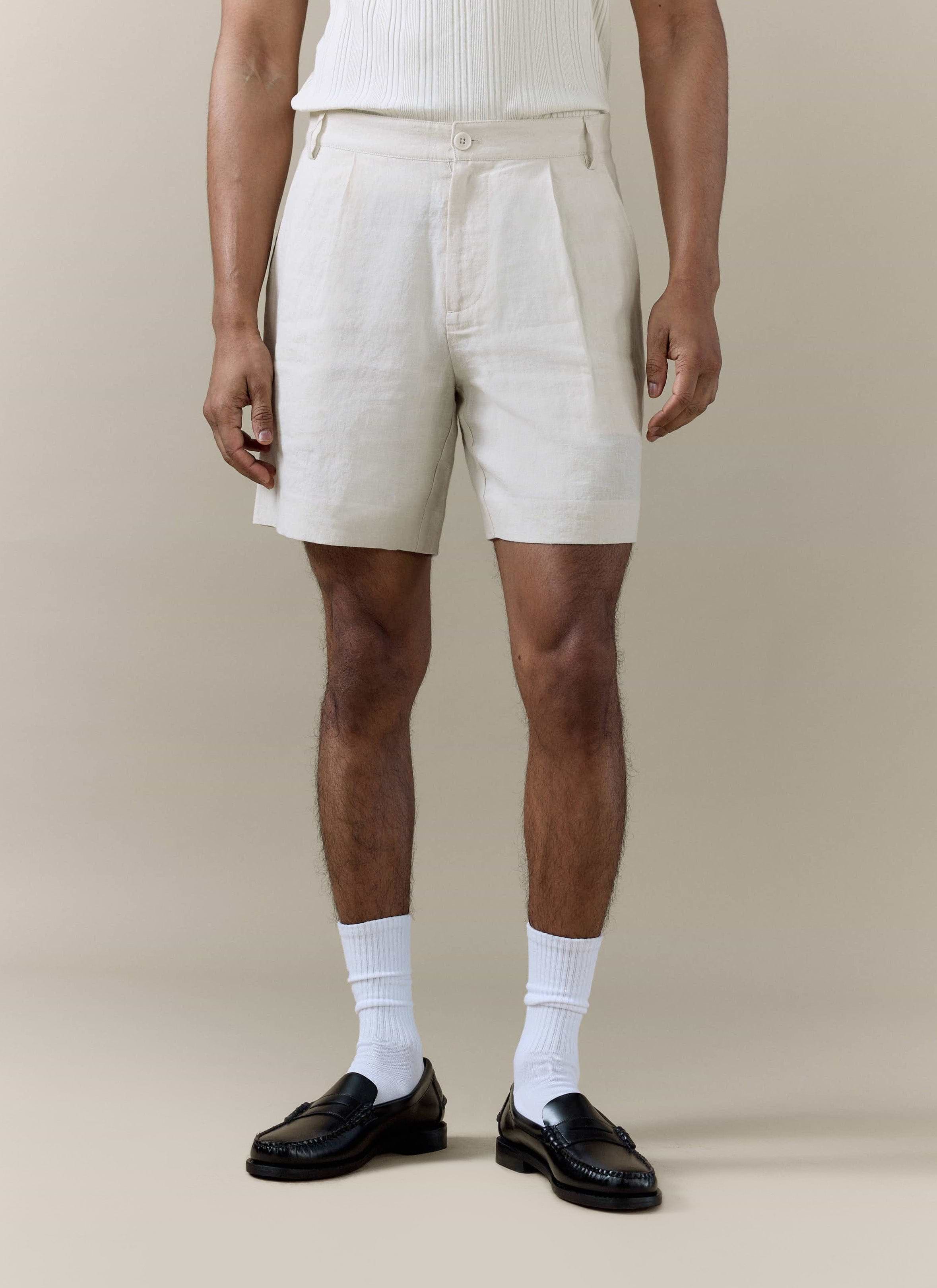 Cream pleated linen shorts by Percival, styled on a model with white socks and black leather loafers.