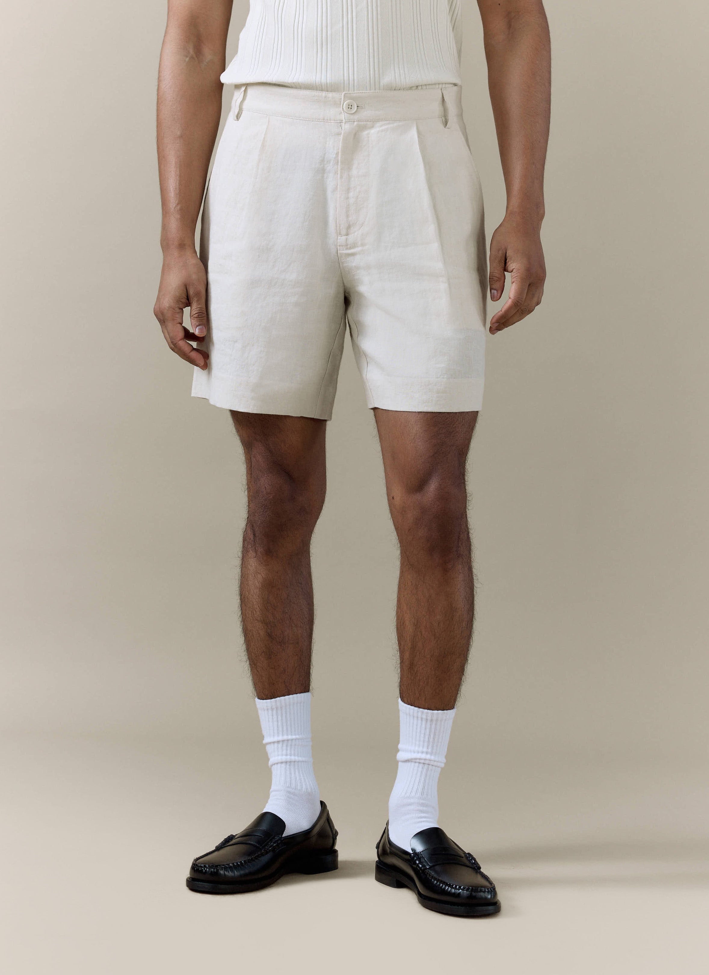 Cream pleated linen shorts by Percival, styled on a model with white socks and black leather loafers.