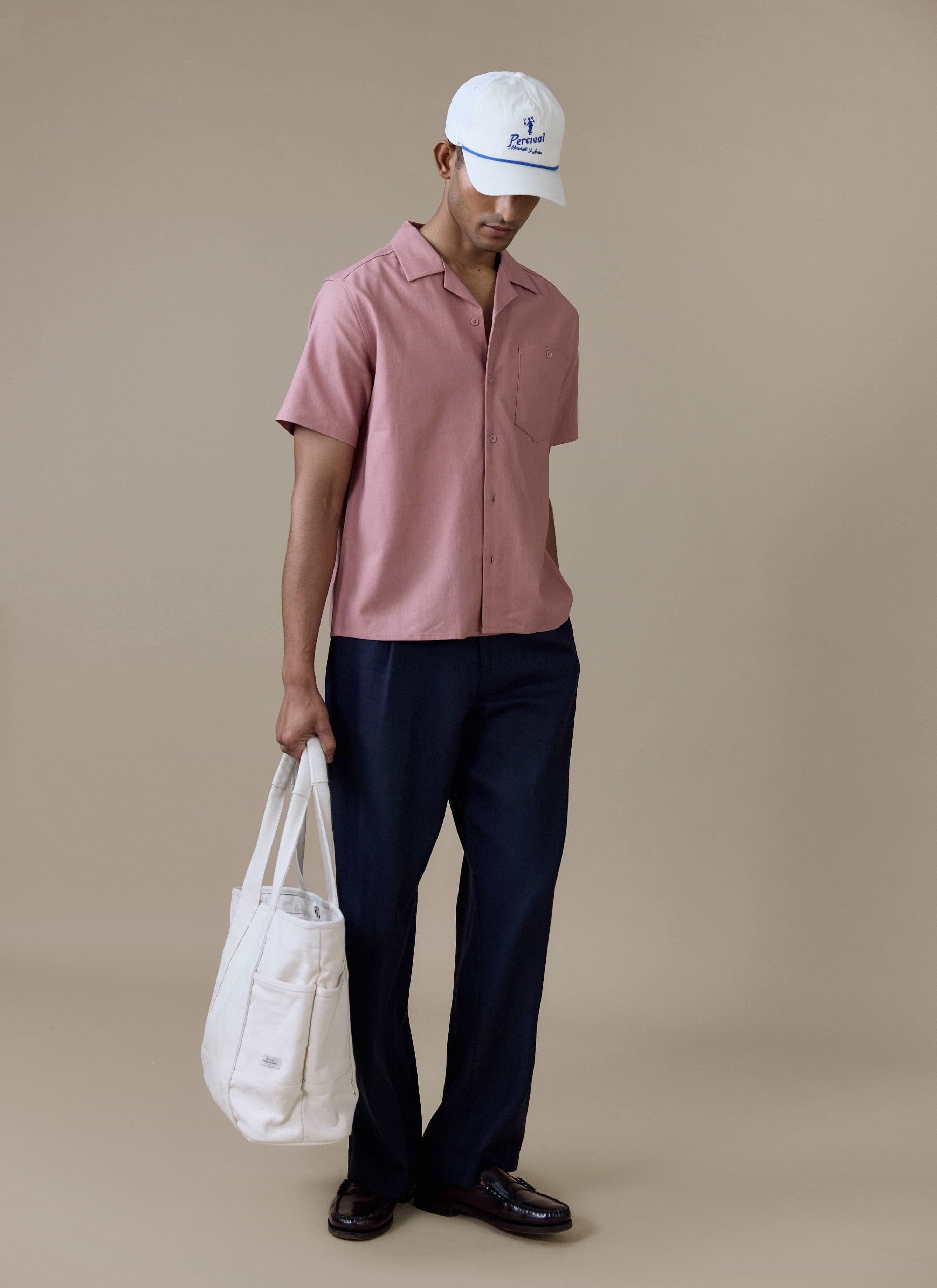 Percival dusty pink linen short sleeve Cuban shirt, styled with navy trousers, a white tote bag and a white cap.