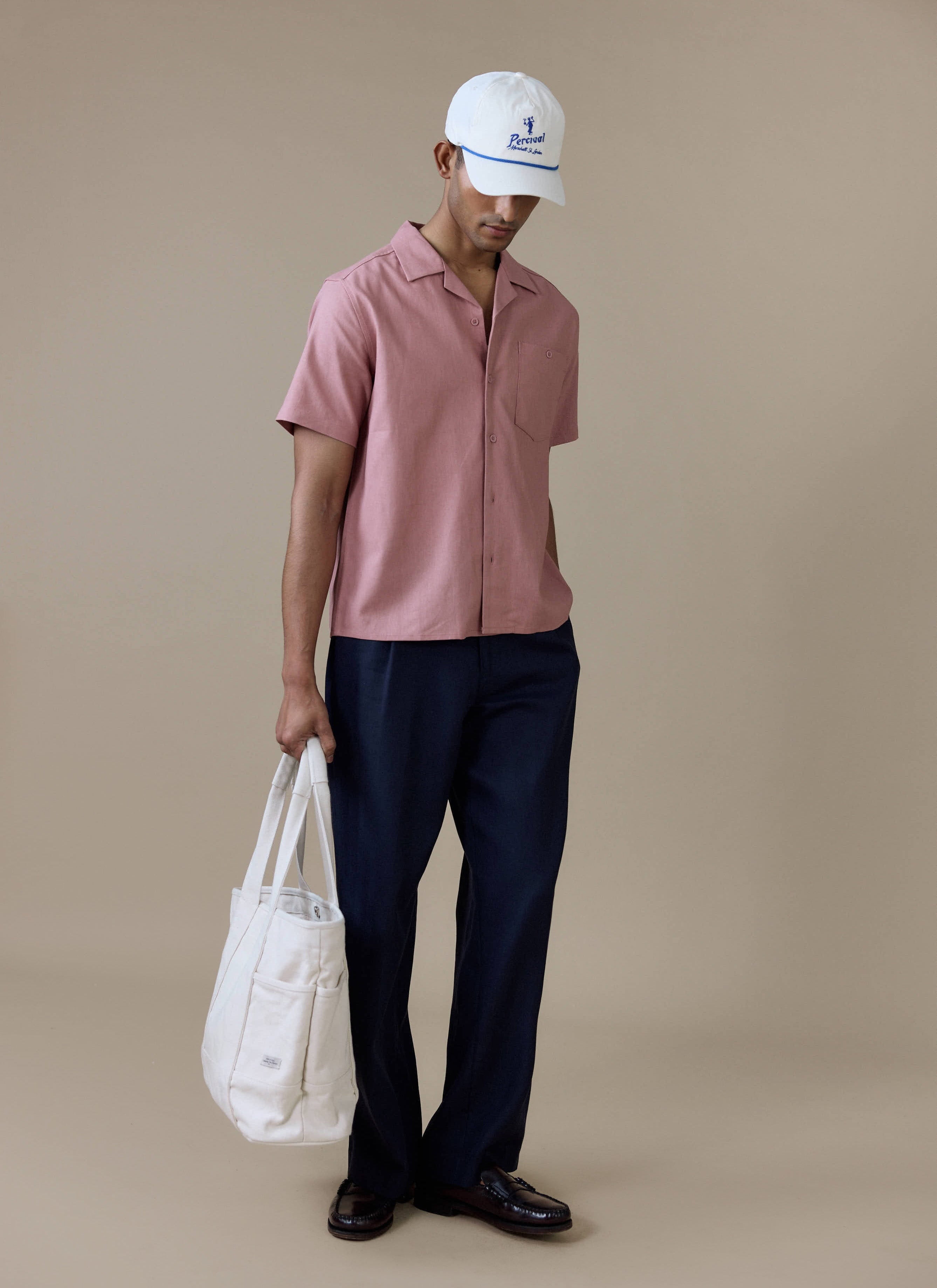 Percival dusty pink linen short sleeve Cuban shirt, styled with navy trousers, a white tote bag and a white cap.