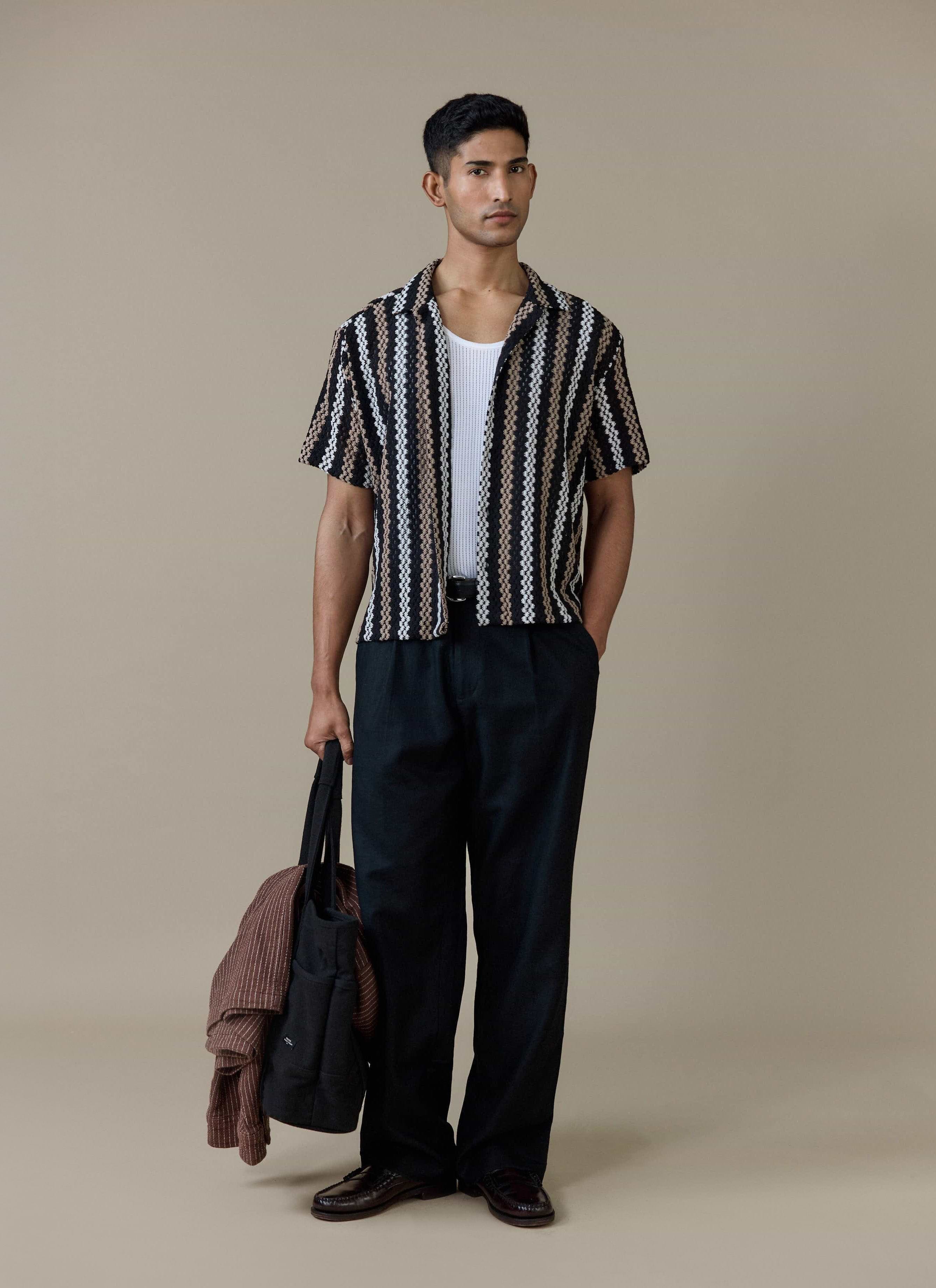 A model wearing the Percival Wade Crochet Boxy Shirt in black stripe over a white vest and black trousers.