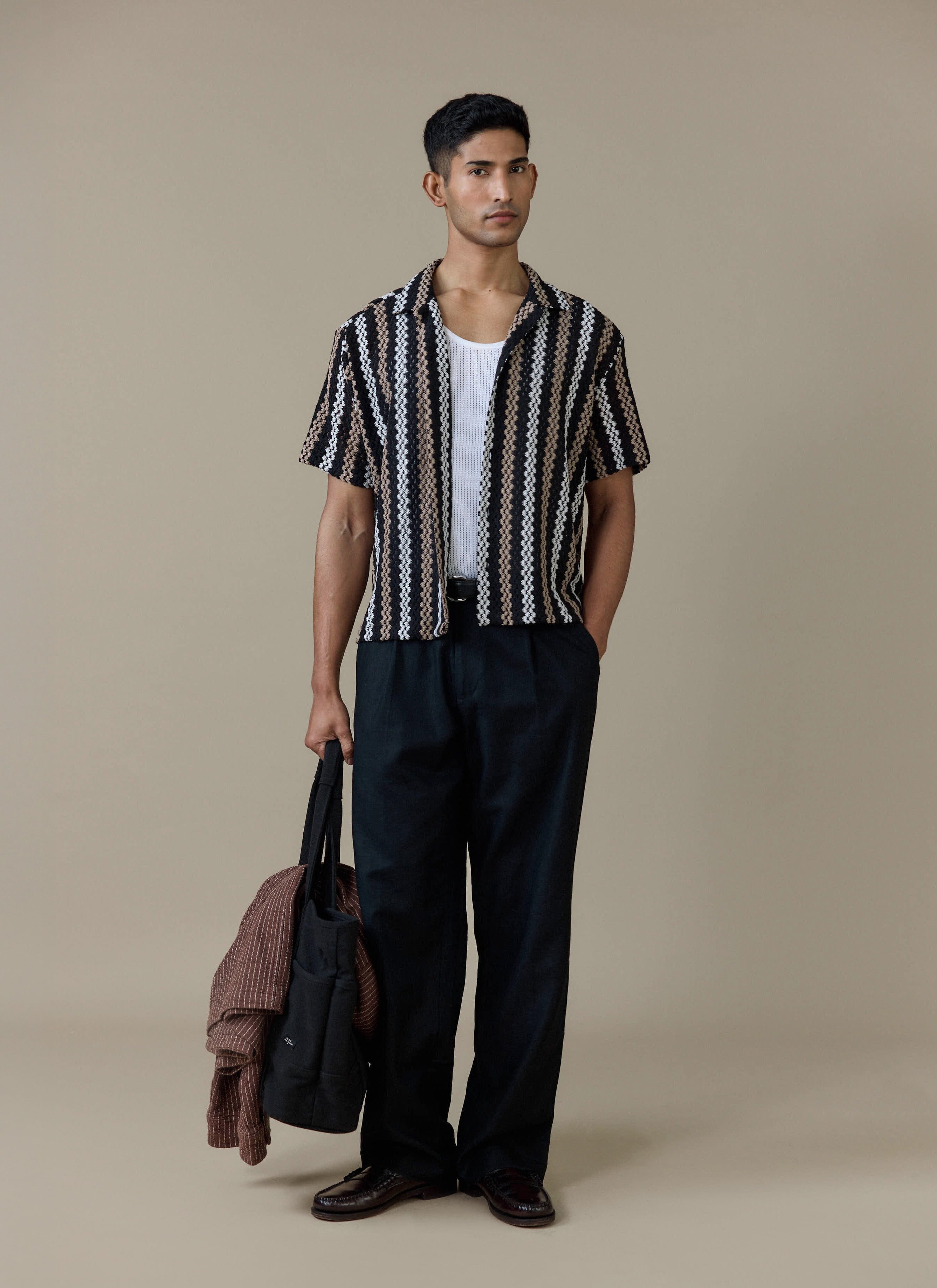 A model wearing the Percival Wade Crochet Boxy Shirt in black stripe over a white vest and black trousers.