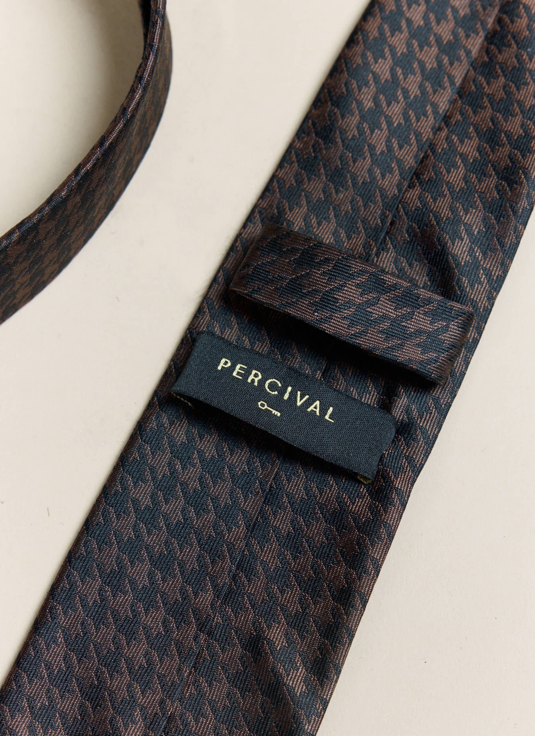 A close-up of the reverse of the Percival brown houndstooth silk tie, showing the brand label and keeper loop.