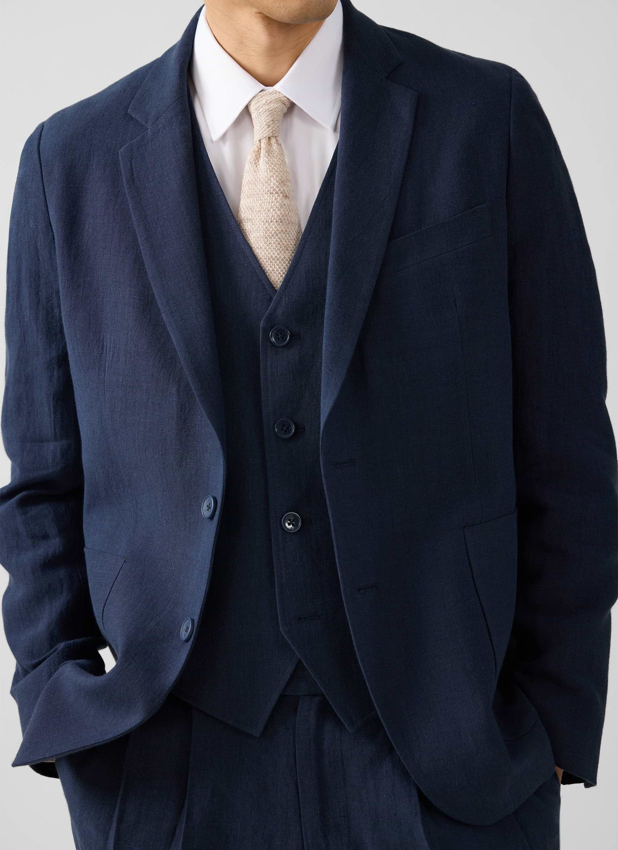 A man models the Percival navy linen waistcoat, styled as part of a three-piece suit with a white shirt and knitted tie.