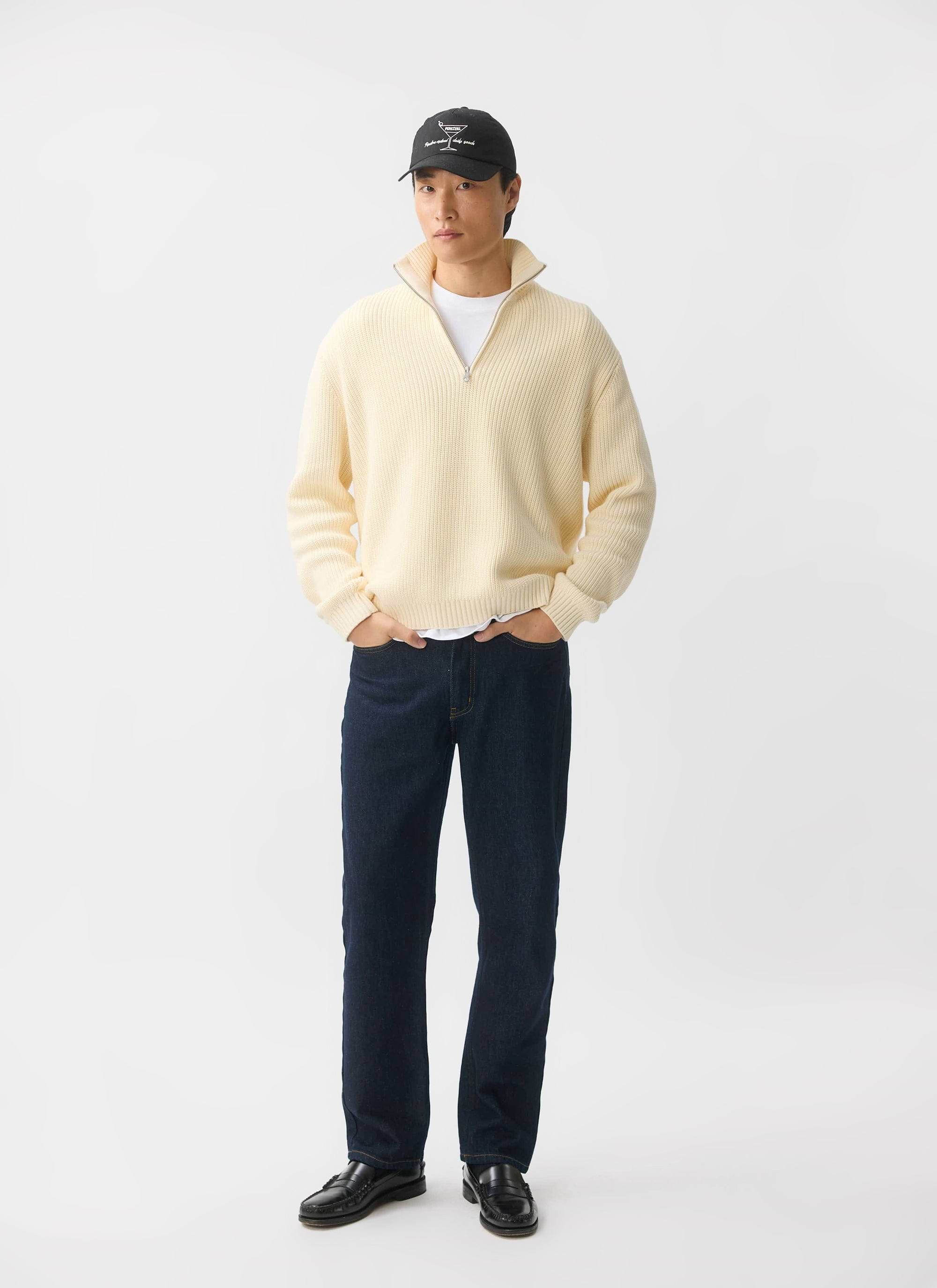 A model wears the ecru Meyer Quarter Zip jumper from Percival, paired with dark wash jeans, loafers and a black baseball cap.