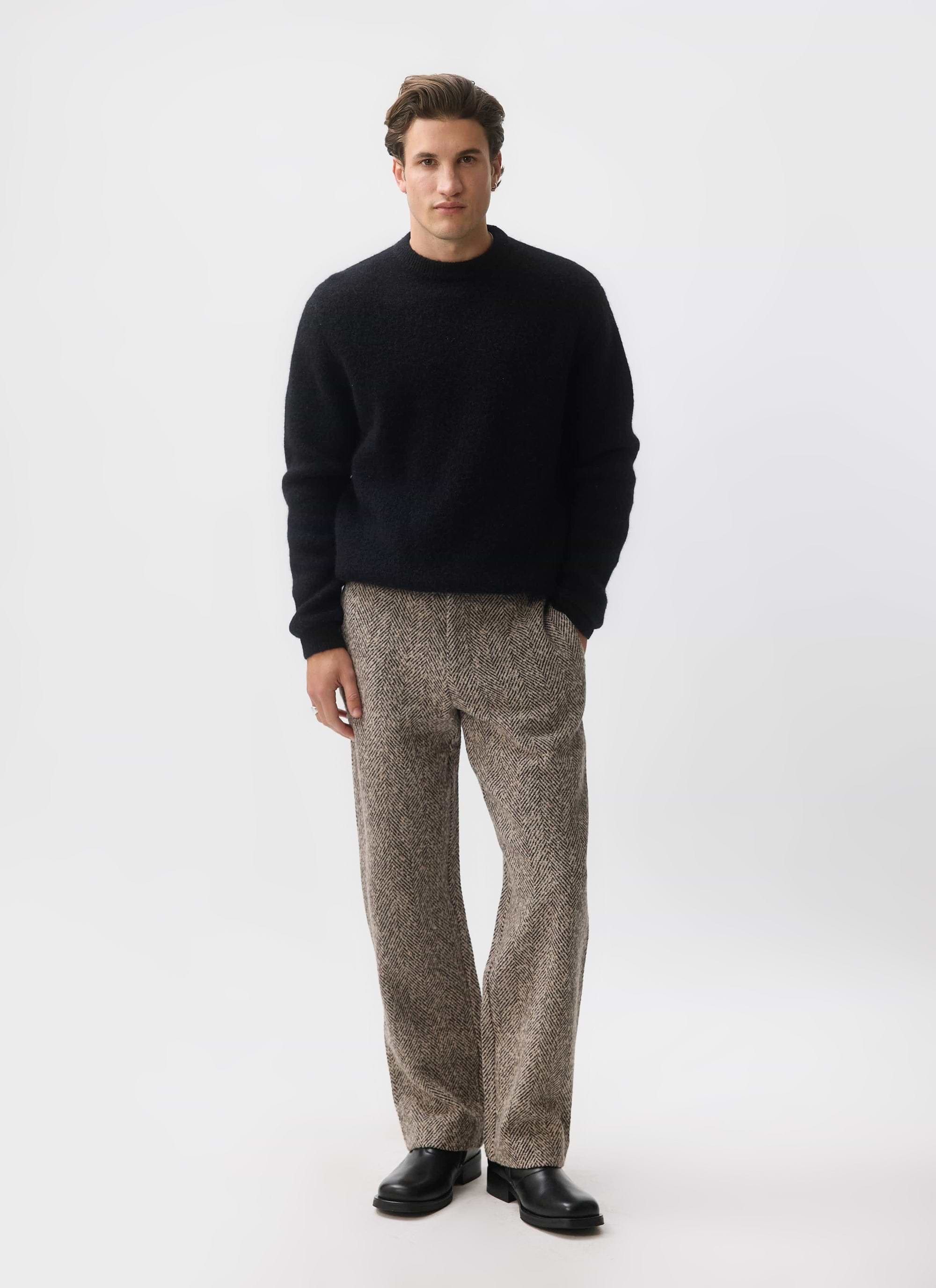 A man models the Percival Herringbone Trousers in ecru with a black jumper and black shoes.