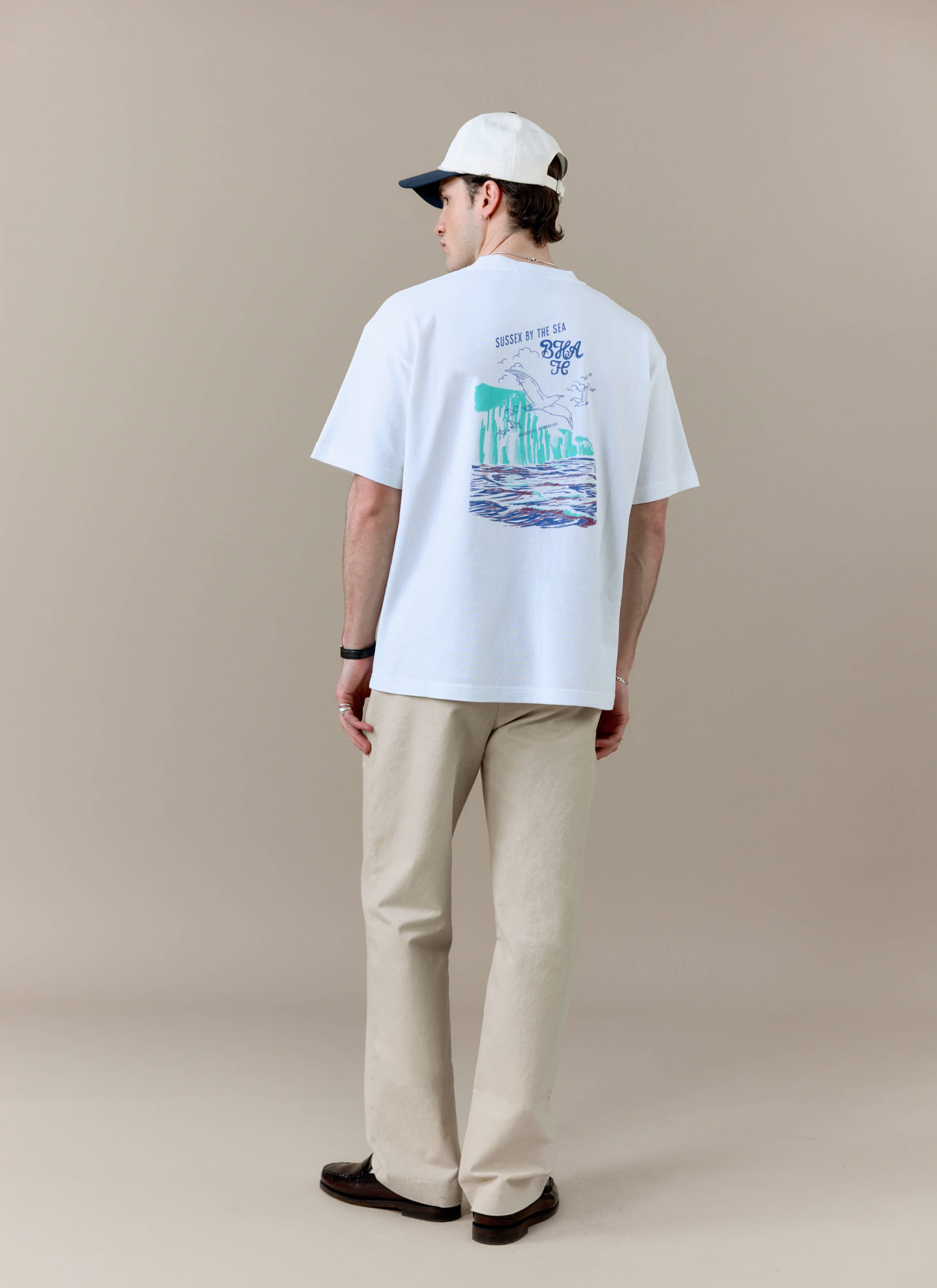 A model from behind wearing the white Percival x Brighton & Hove Albion F.C. t-shirt, showing the colourful back print.