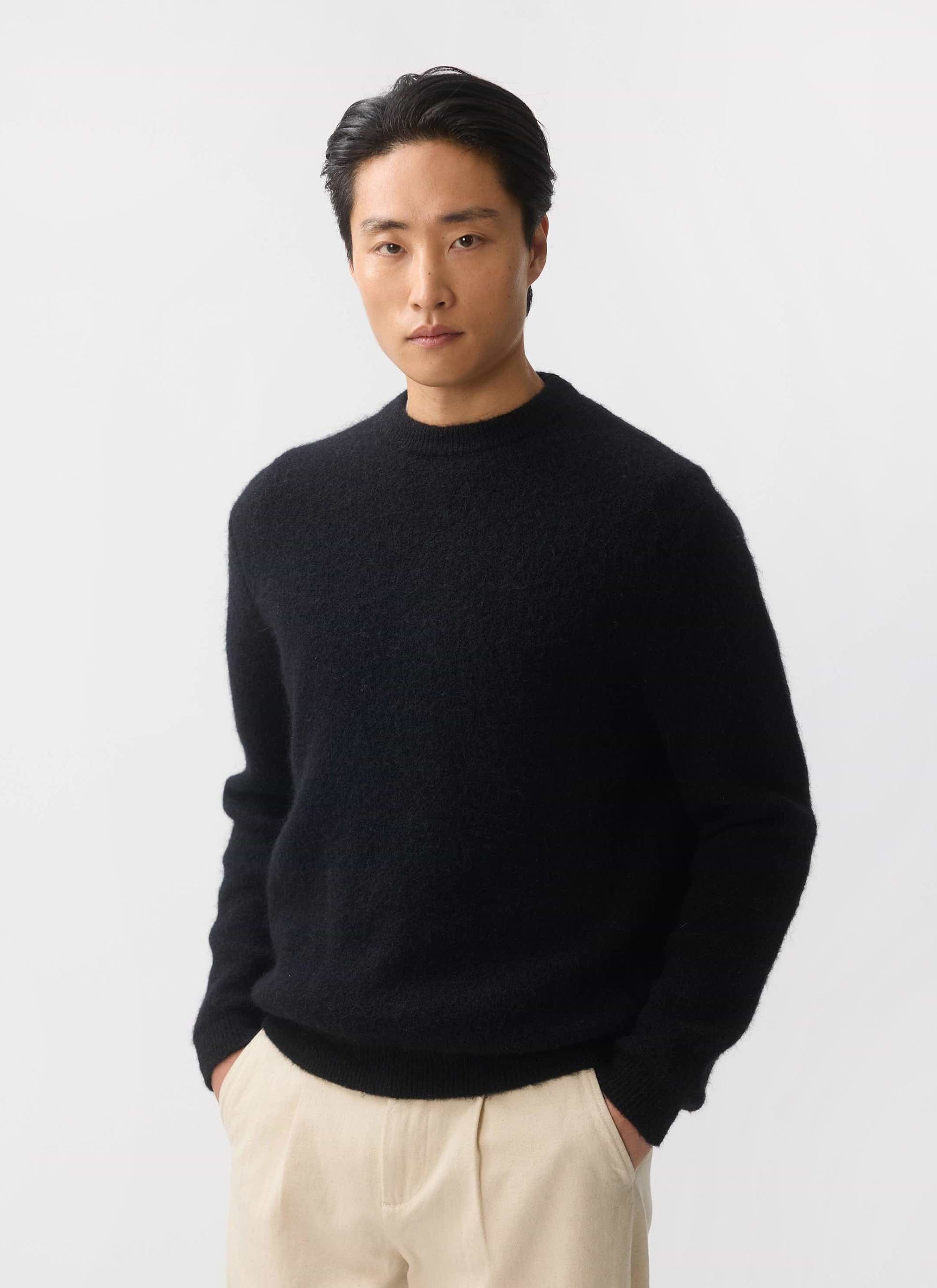 A man models the Percival White Glove crew neck jumper in black mohair, styled with cream trousers against a white background