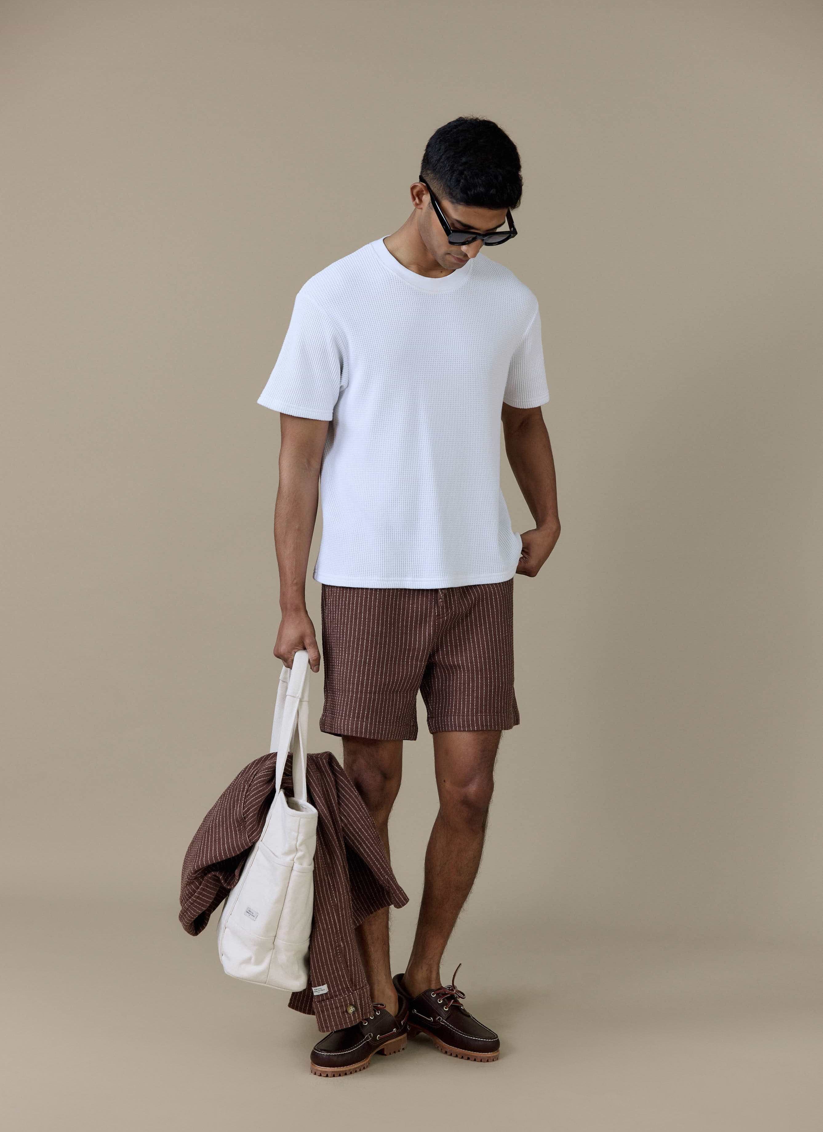 Model wearing the Percival Sorrento Shorts in brown pinstripe linen, styled with a white t-shirt and tote bag.