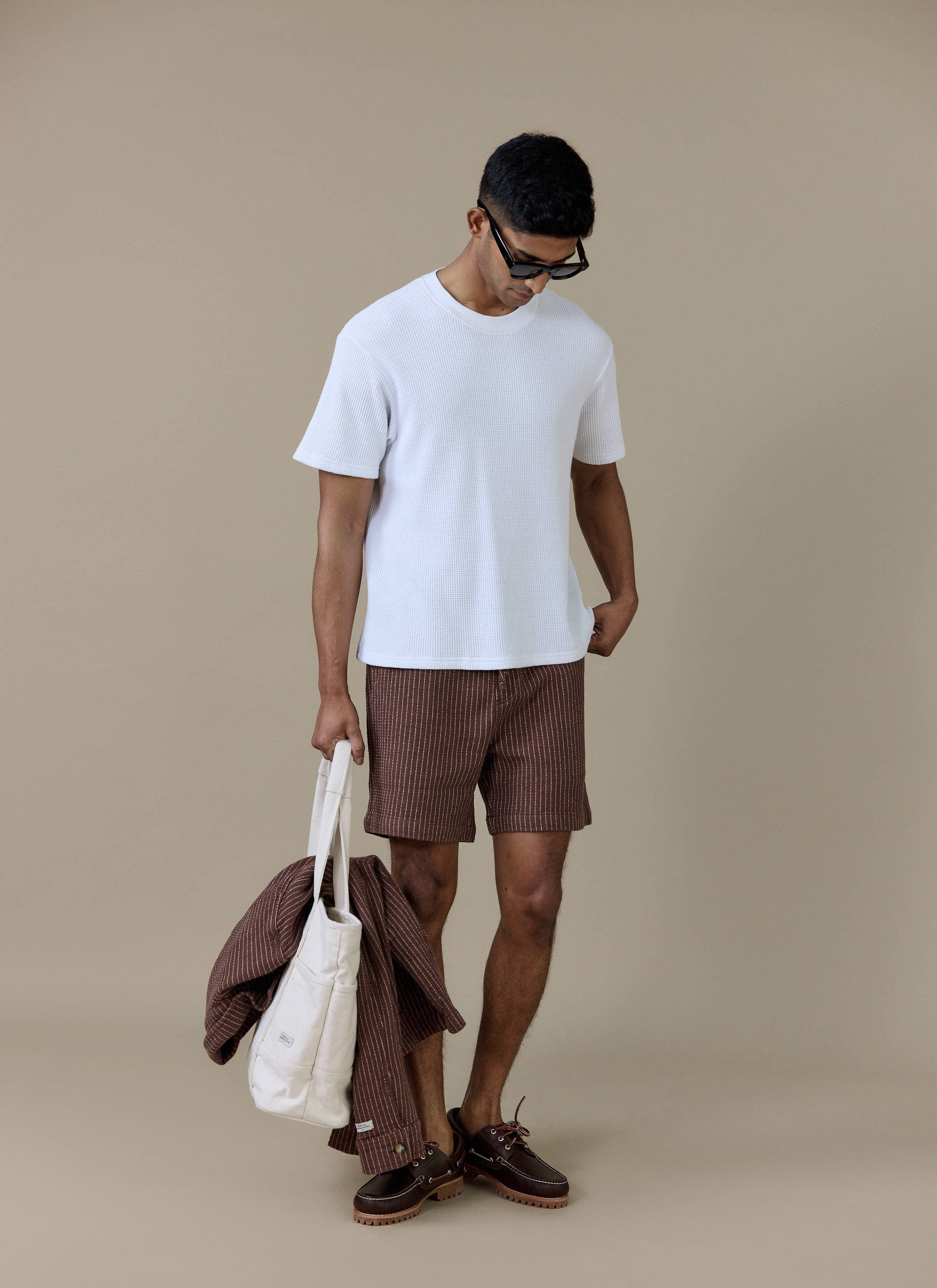 Model wearing the Percival Sorrento Shorts in brown pinstripe linen, styled with a white t-shirt and tote bag.
