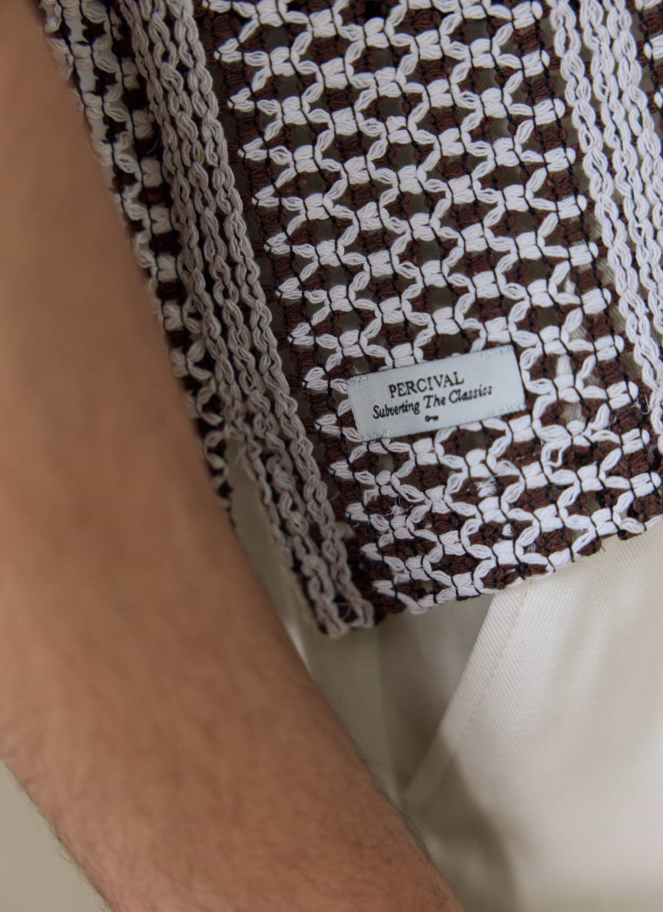 A close-up of the brown crochet knit and Percival 'Subverting The Classics' label on the Hugh Crochet Boxy Shirt.