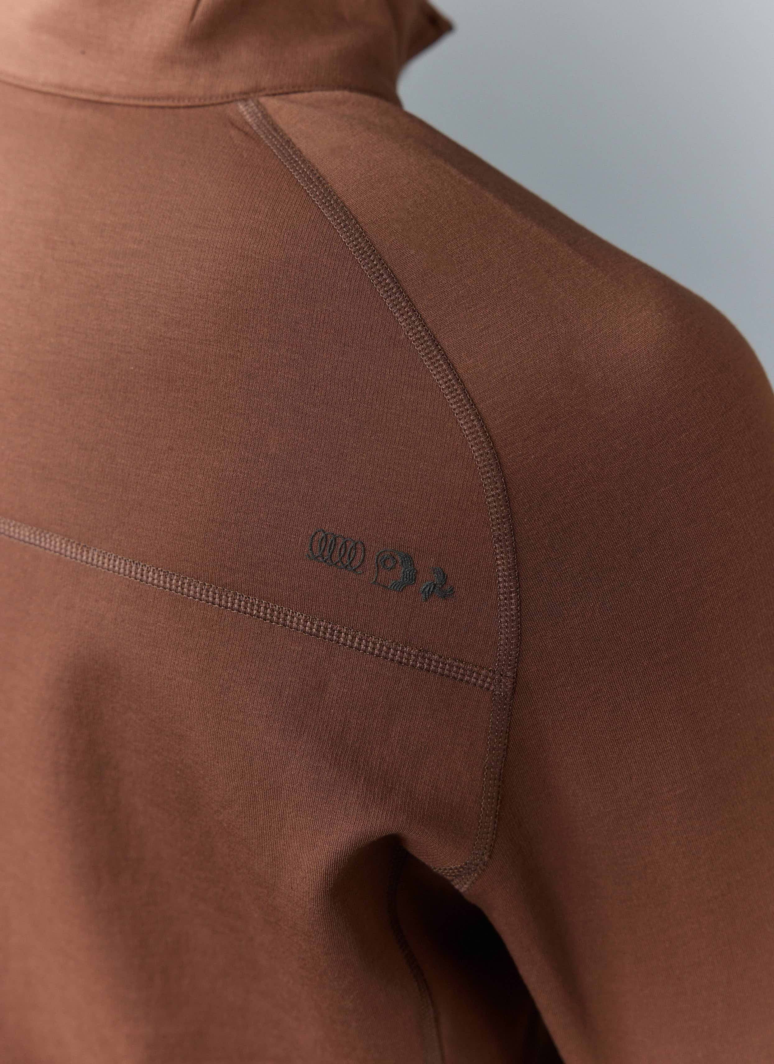 A close-up of the Percival PCVL Base Layer Quarter Zip Jacket in rust, showing the chest logo and raglan sleeve detail.