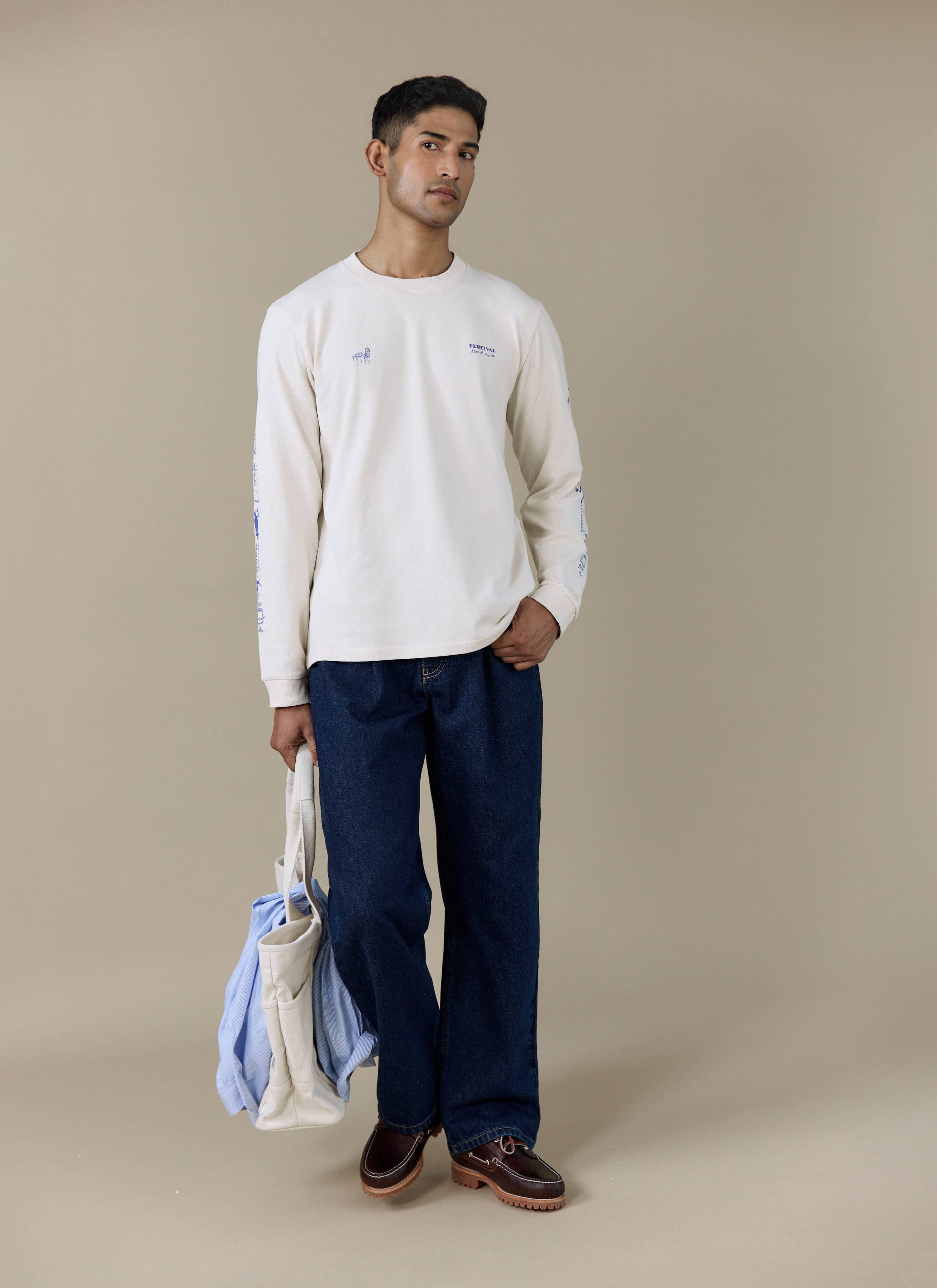 A model wears the Percival Harlow Fine Wines Long Sleeve T-Shirt in cream, featuring blue graphic prints on the sleeves.