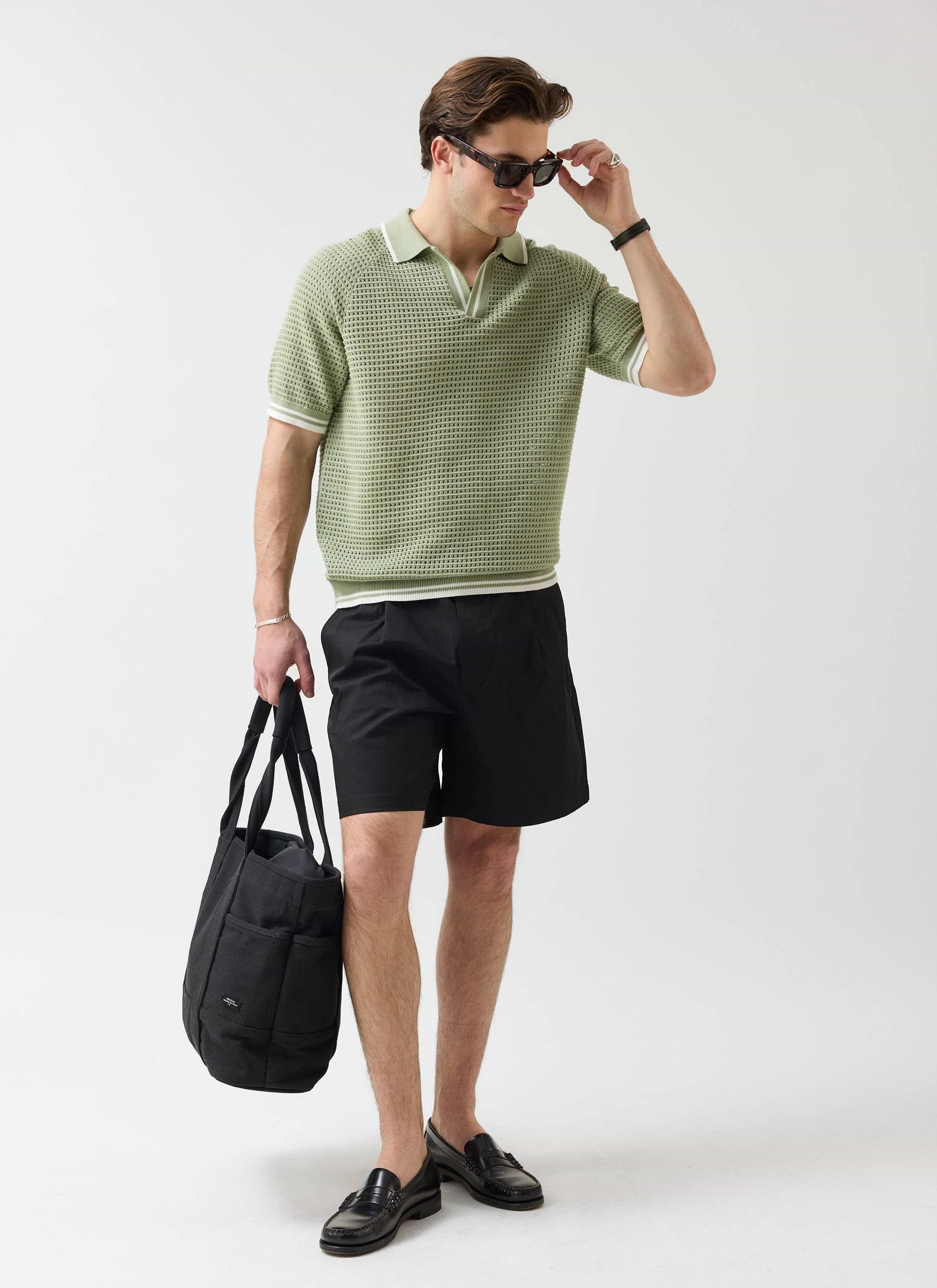 A model wearing the Percival sage knitted tennis polo, black shorts, and sunglasses, holding a large black tote bag.