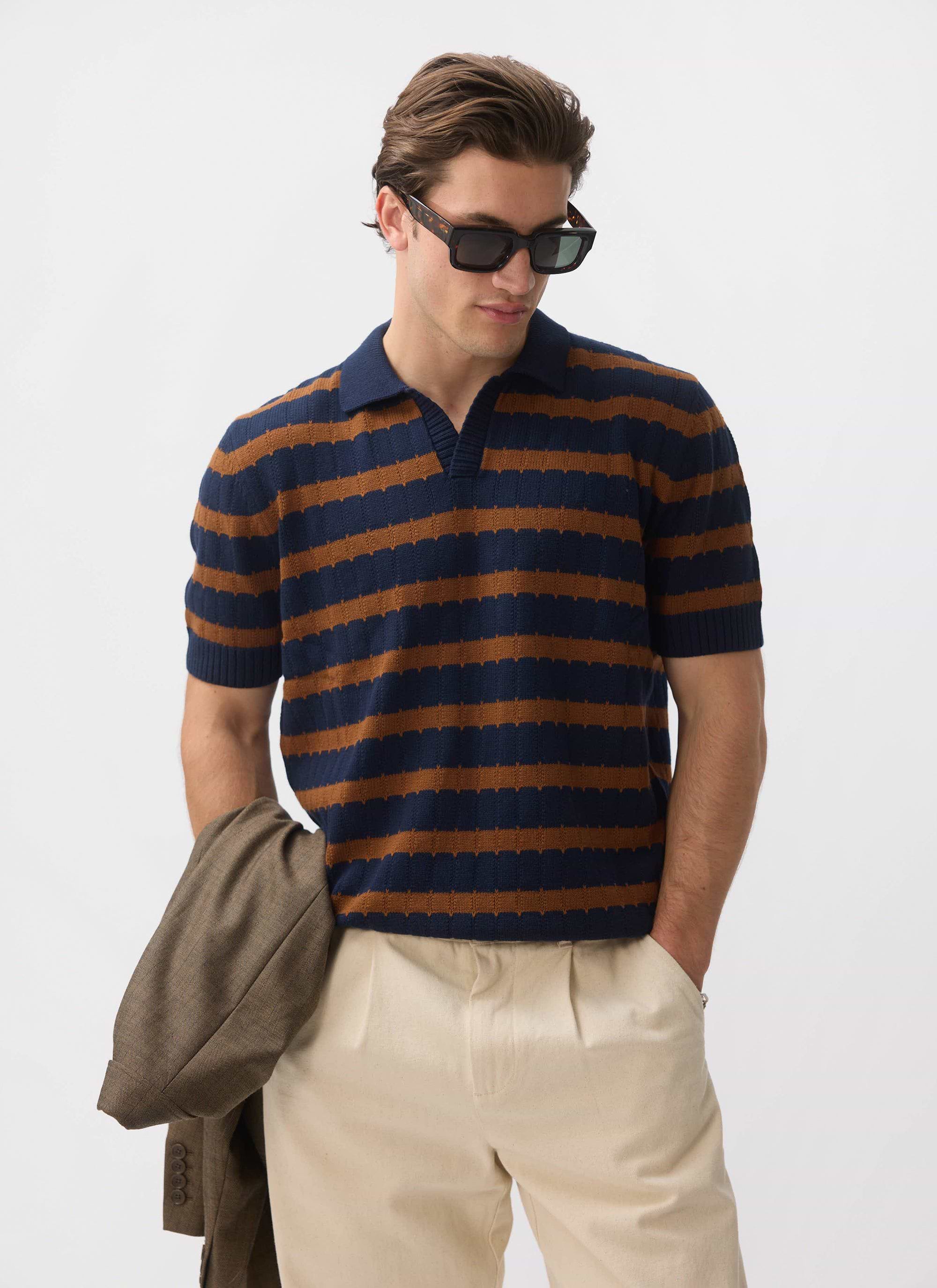 A man models the Percival Stine Knitted Polo in brown and navy, styled with cream trousers and holding a brown blazer.