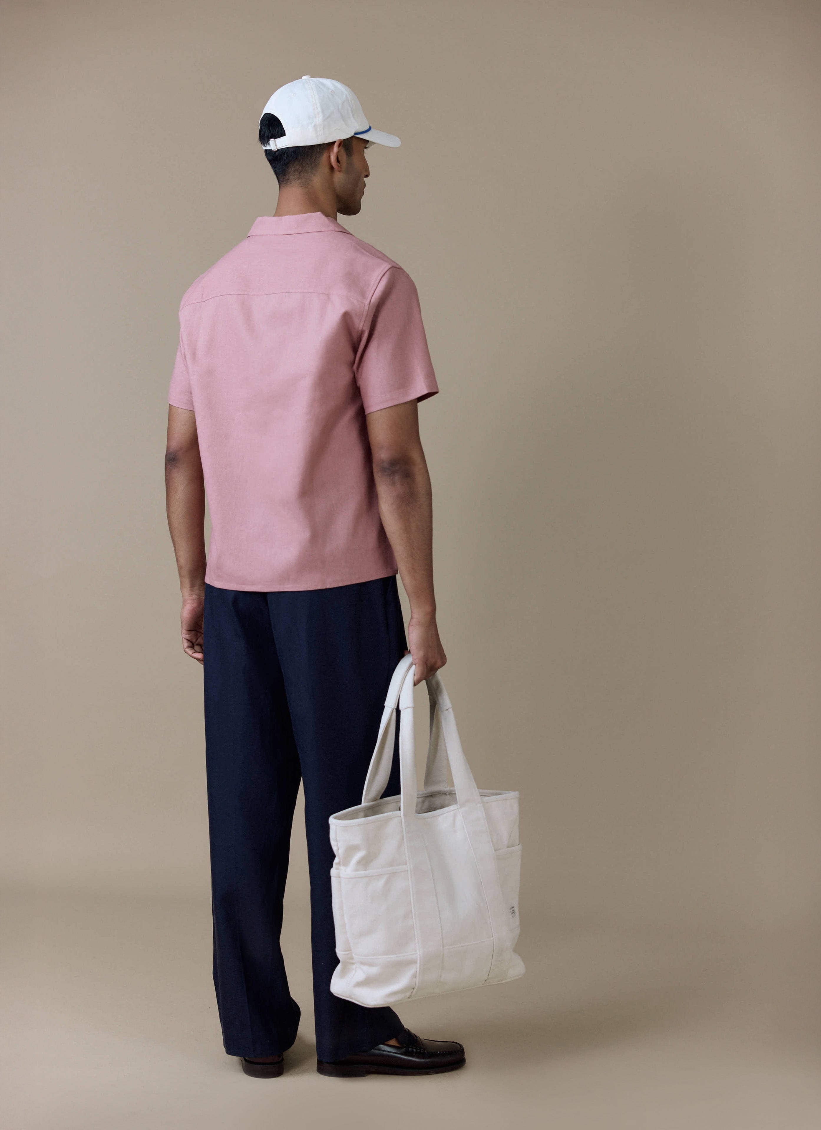 Dusty pink Percival linen Short Sleeve Cuban Shirt, shown from the back with dark trousers and a large white tote bag.