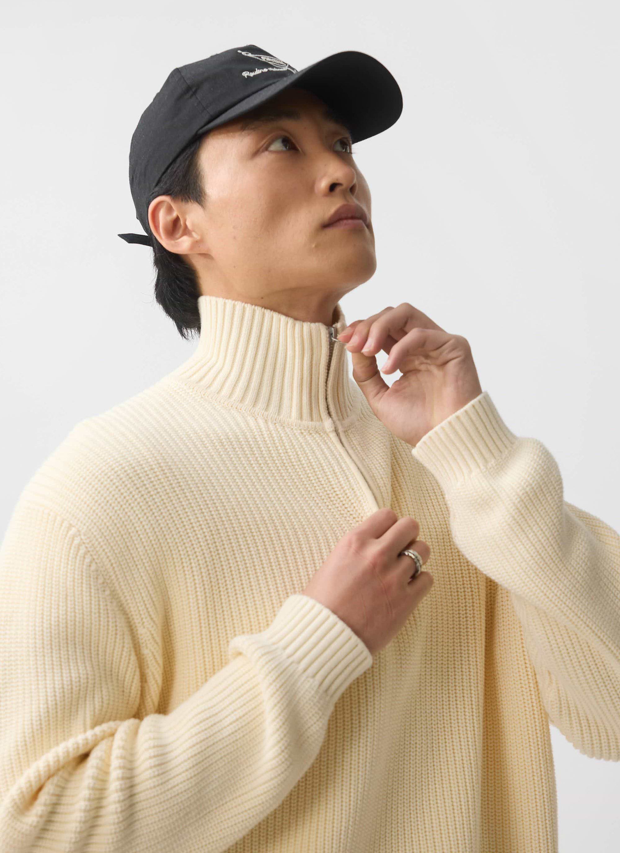A model in a black cap adjusts the zip on the ecru Meyer Quarter Zip Fisherman knit jumper from Percival.