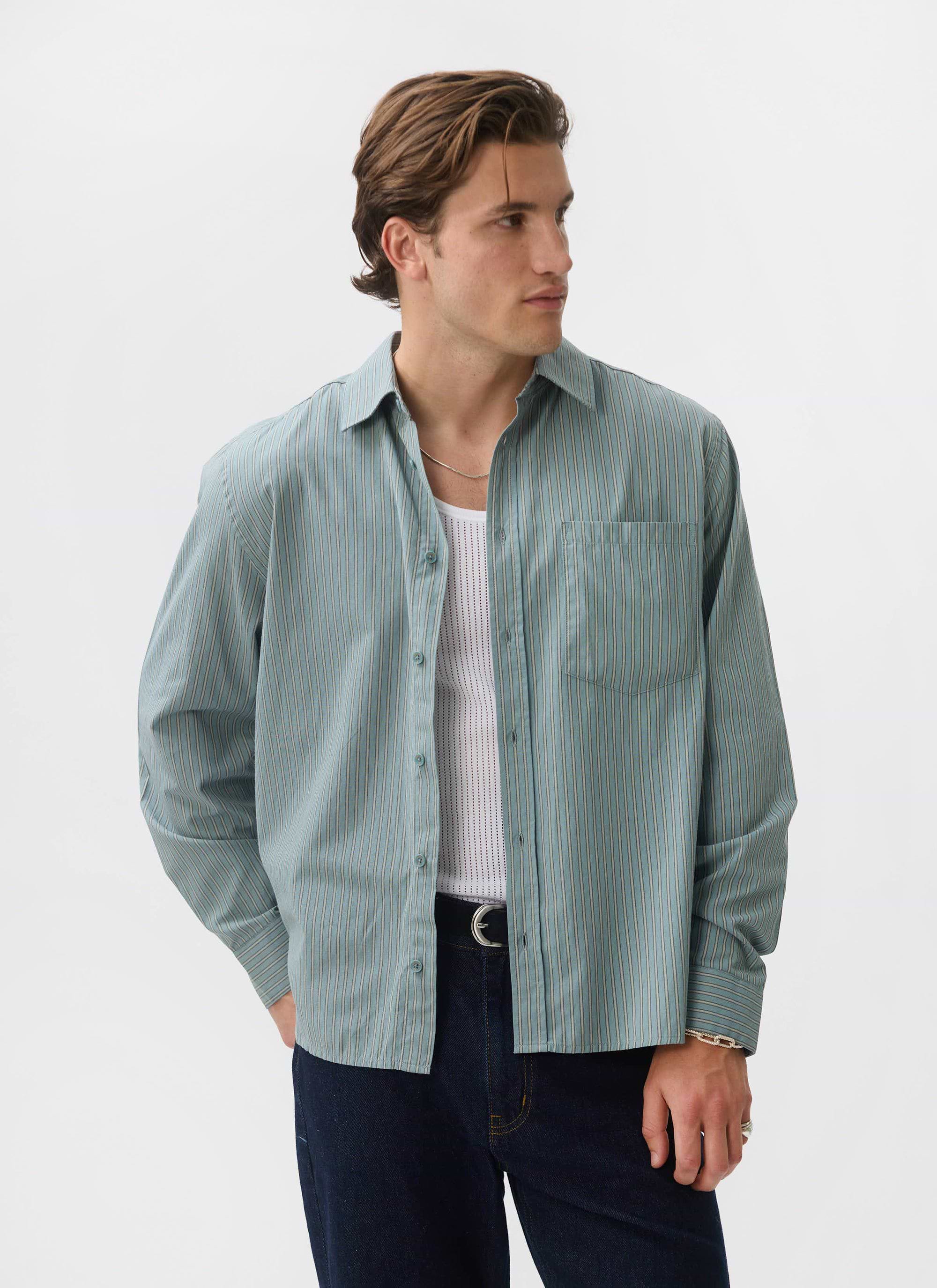 A man models the Percival Clancy Striped Long Sleeve Shirt in blue, worn open over a white vest and dark jeans.