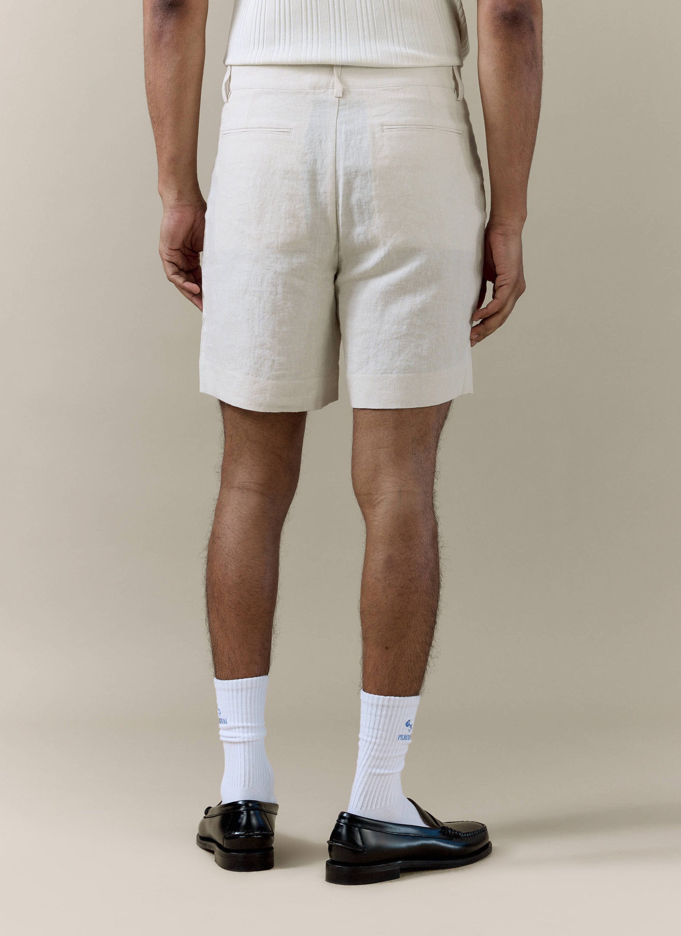 Cream pleated linen shorts by Percival, shown from the back with welt pocket details and a relaxed summer fit.