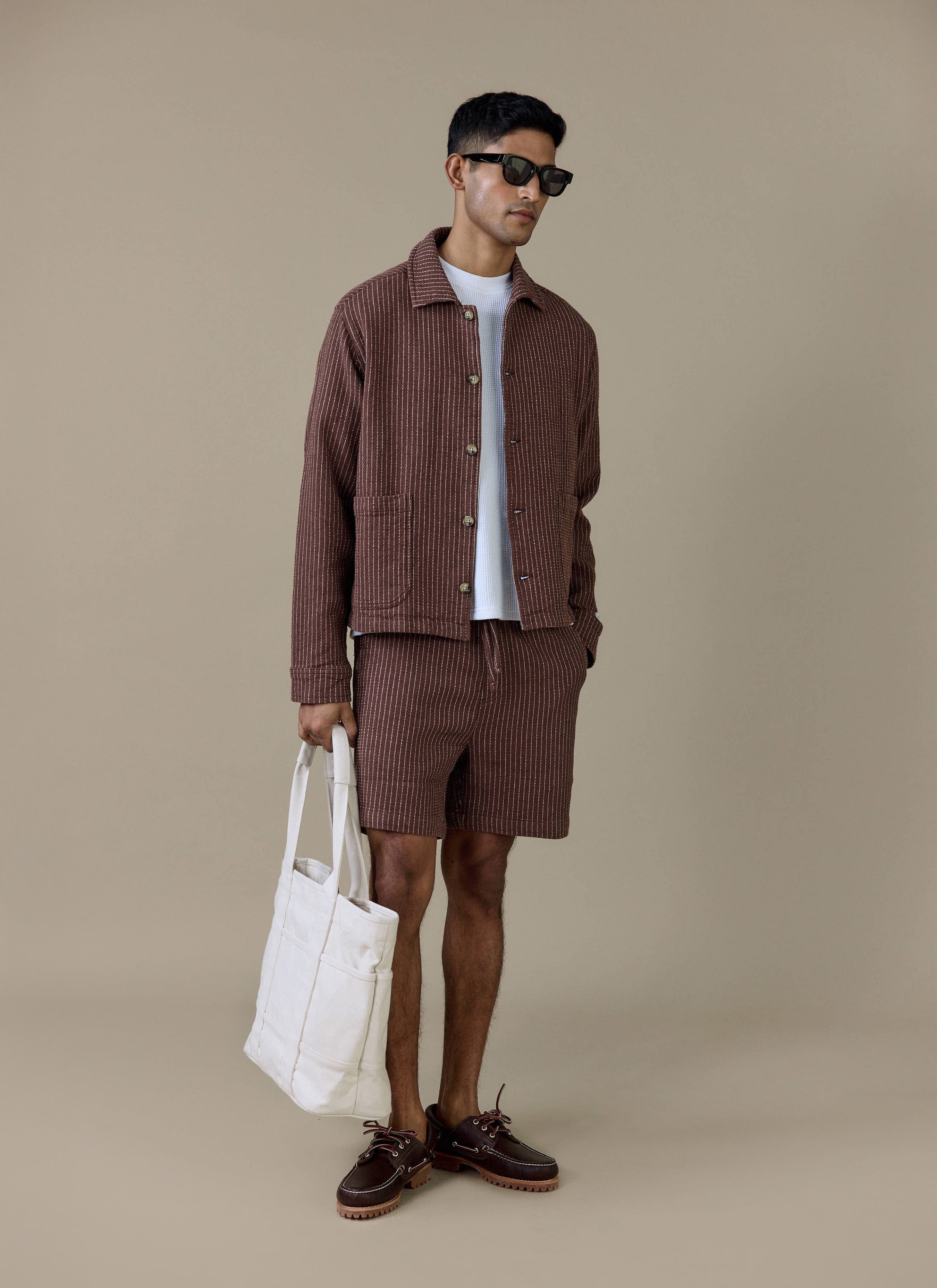 Model wears the Percival Sorrento Shorts in brown linen with a matching pinstripe jacket and white tote bag.