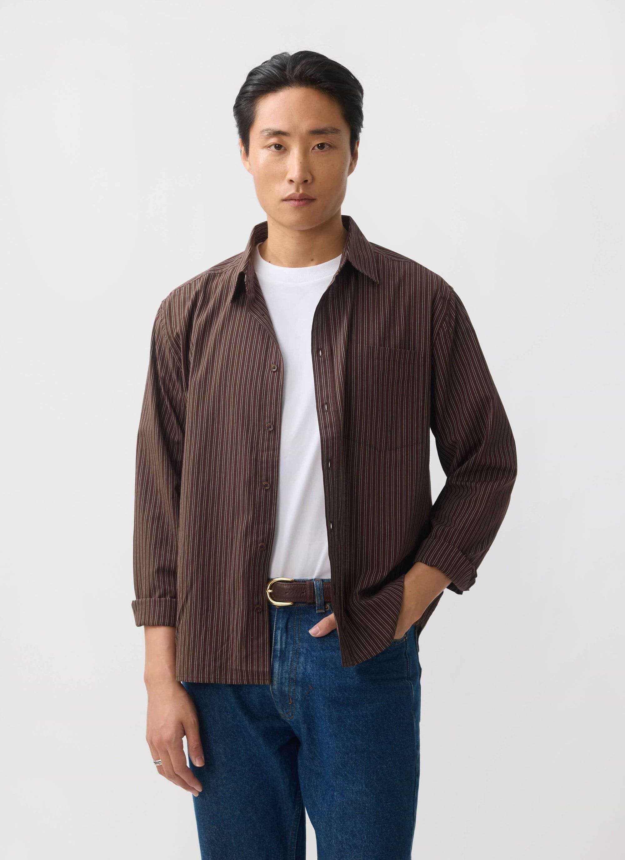A model wears the Percival Clancy Striped Long Sleeve Shirt in brown, styled open over a white t-shirt and blue jeans.