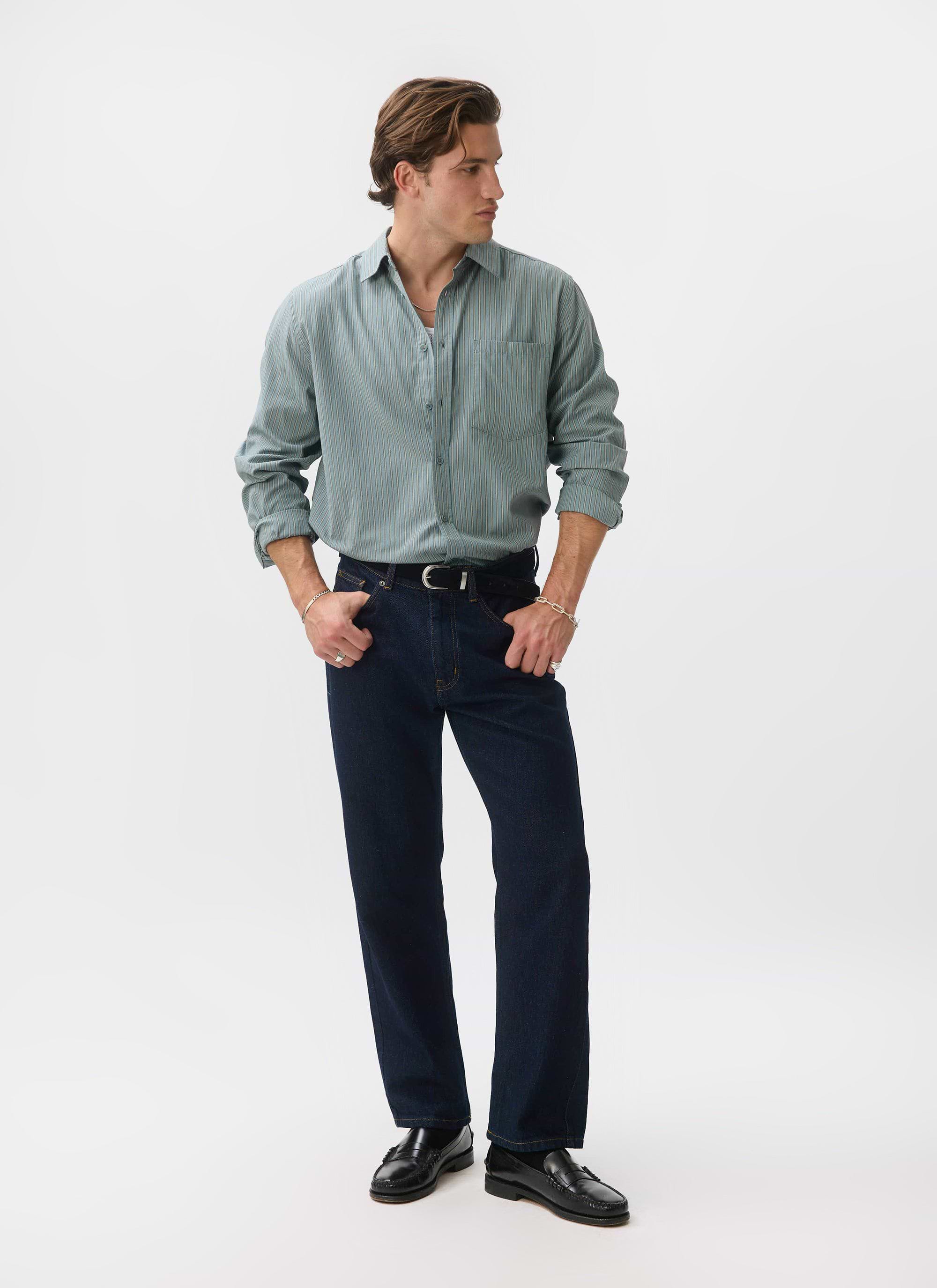 A man models the Percival Clancy Striped Long Sleeve Shirt in blue, styled with dark denim jeans and black loafers.