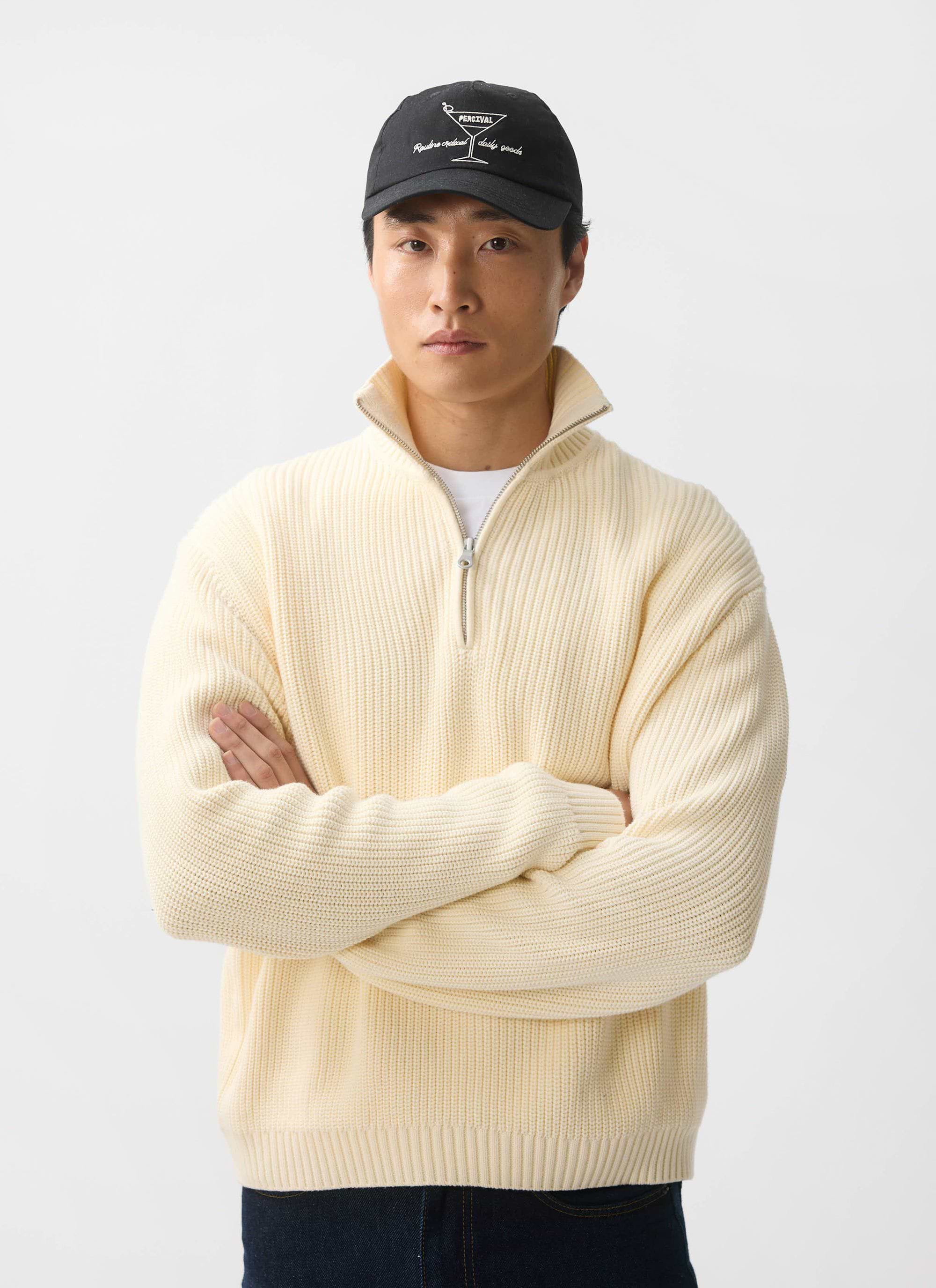 A model wears the Percival Meyer Quarter Zip Fisherman jumper in ecru, styled with a black cap against a white background.