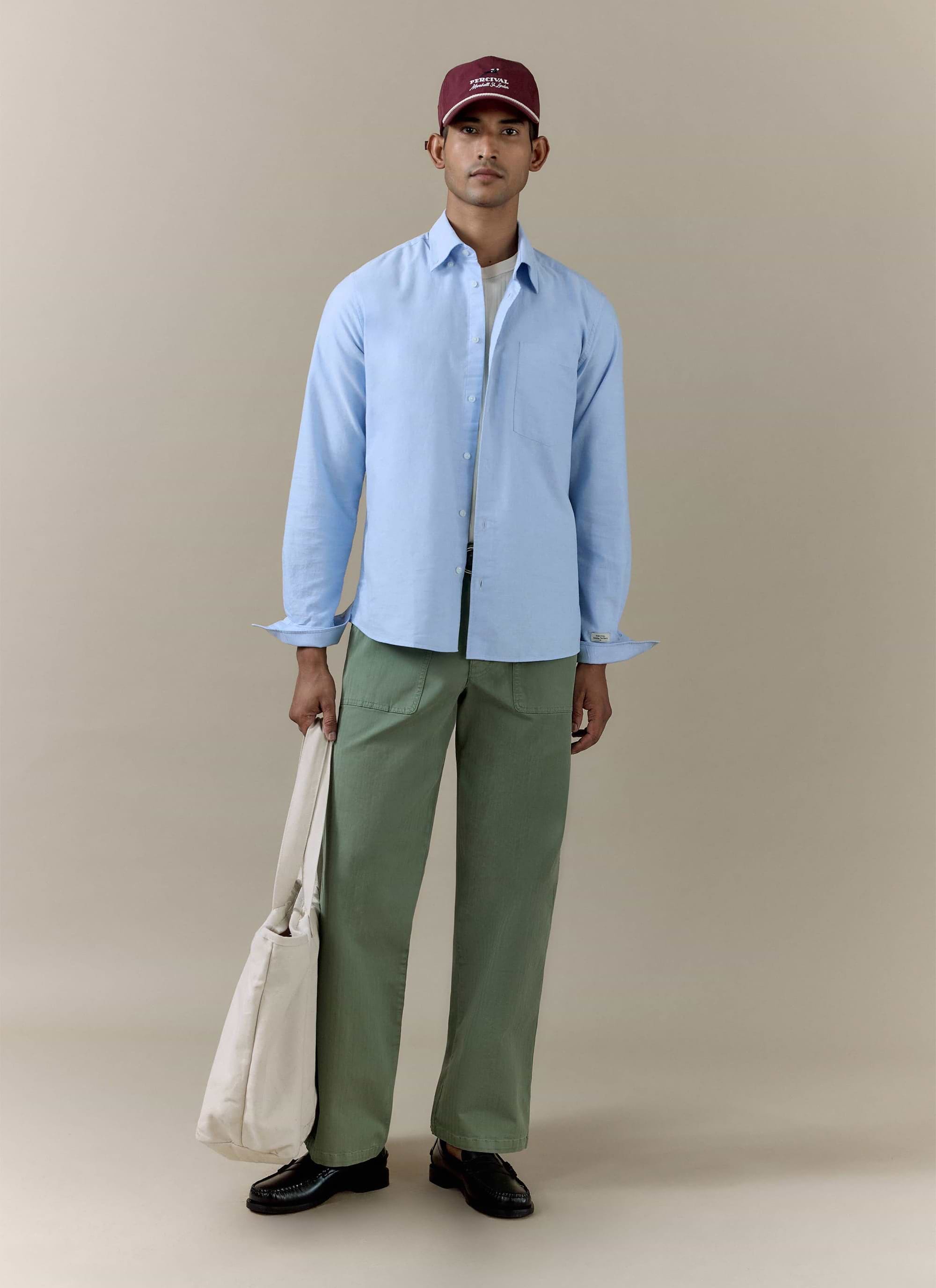 Model wearing the sage green cotton Lungo Herringbone Straight Trousers by Percival with a blue shirt and tote bag.