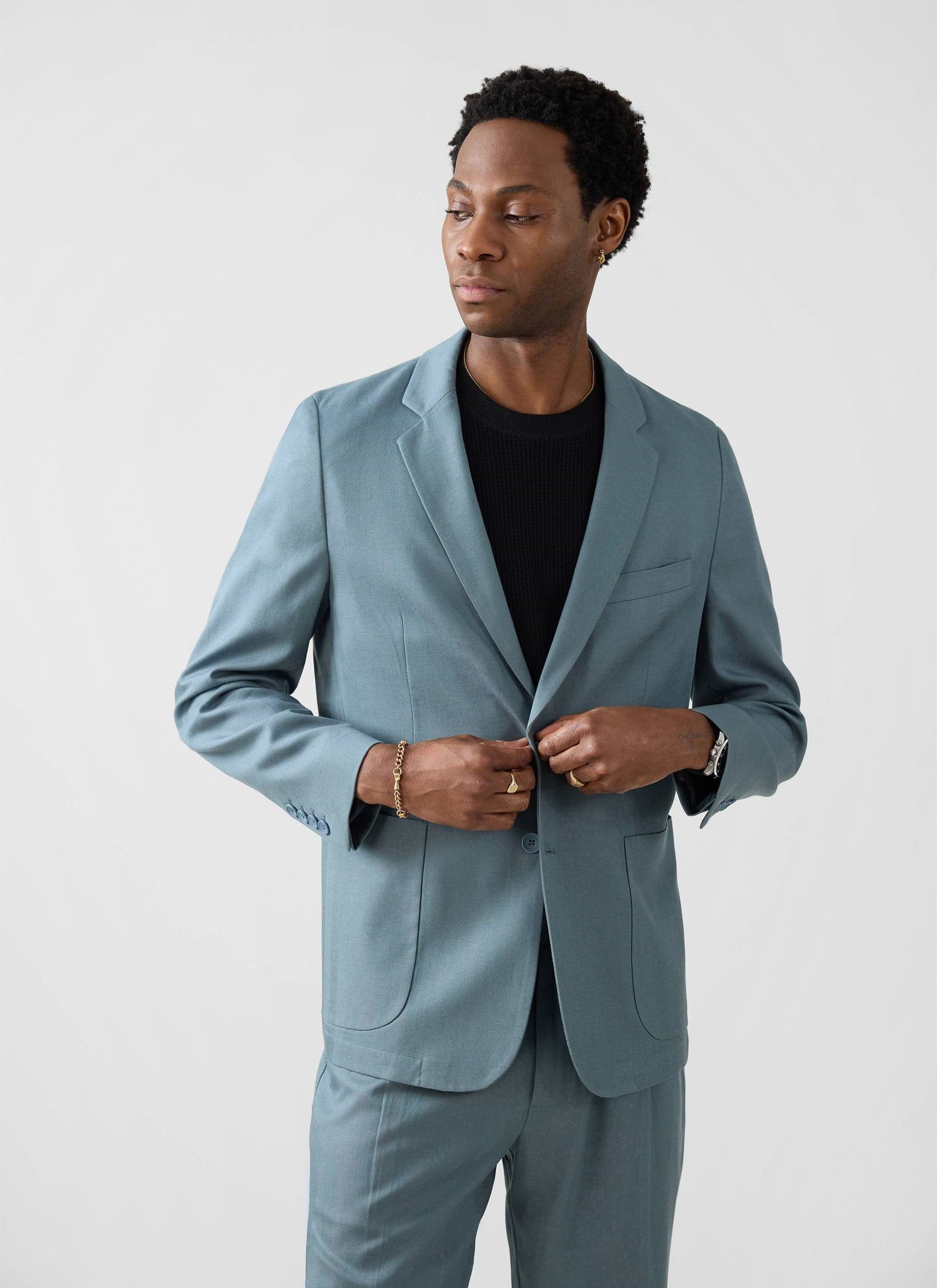 A man models the Percival Lyocell Regular Blazer in Slate Blue, buttoning the jacket against a plain white background.