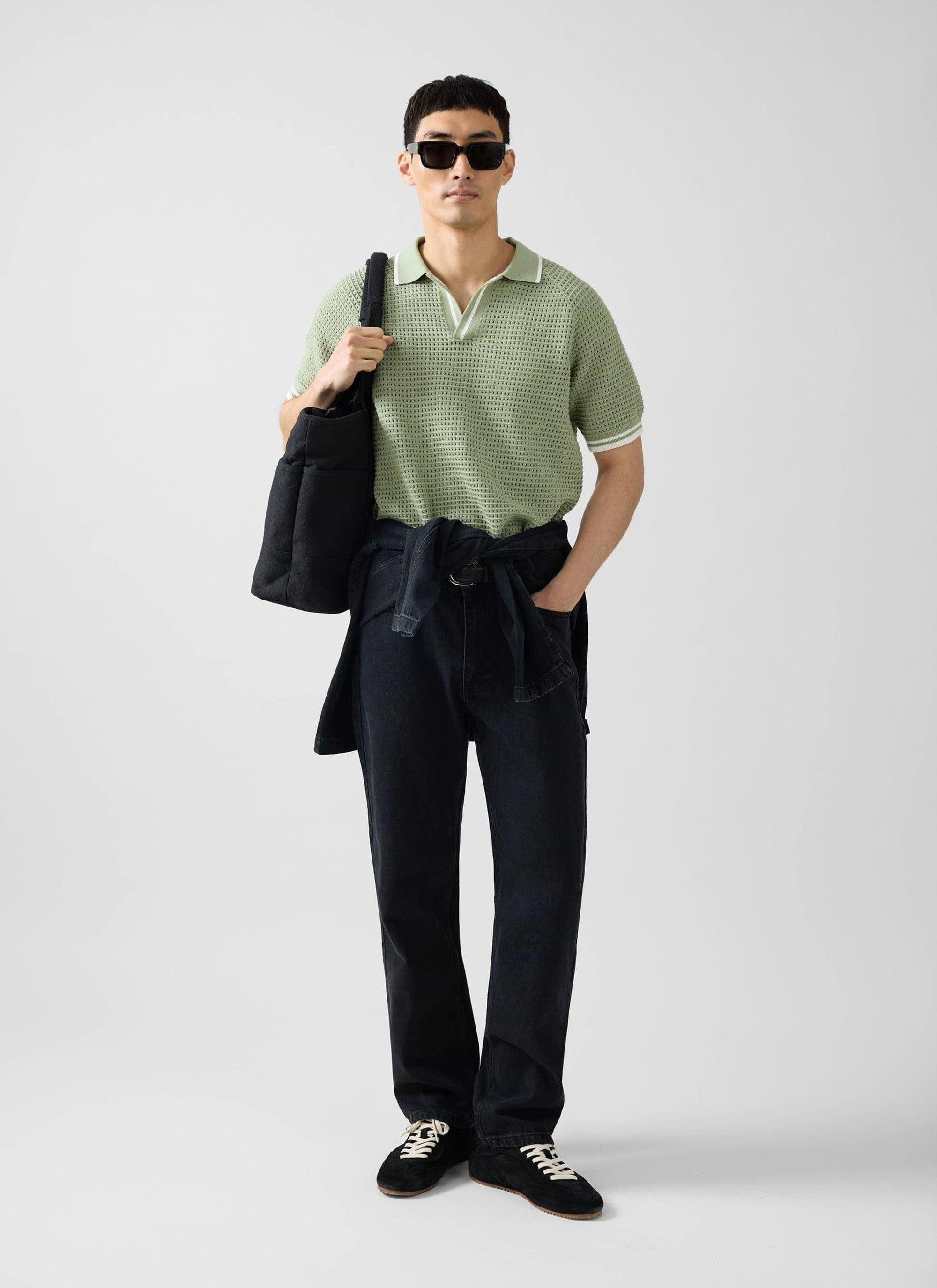 A man wearing the Percival sage green Tennis Boxy Knitted Polo, black jeans, sunglasses, and carrying a black tote bag.