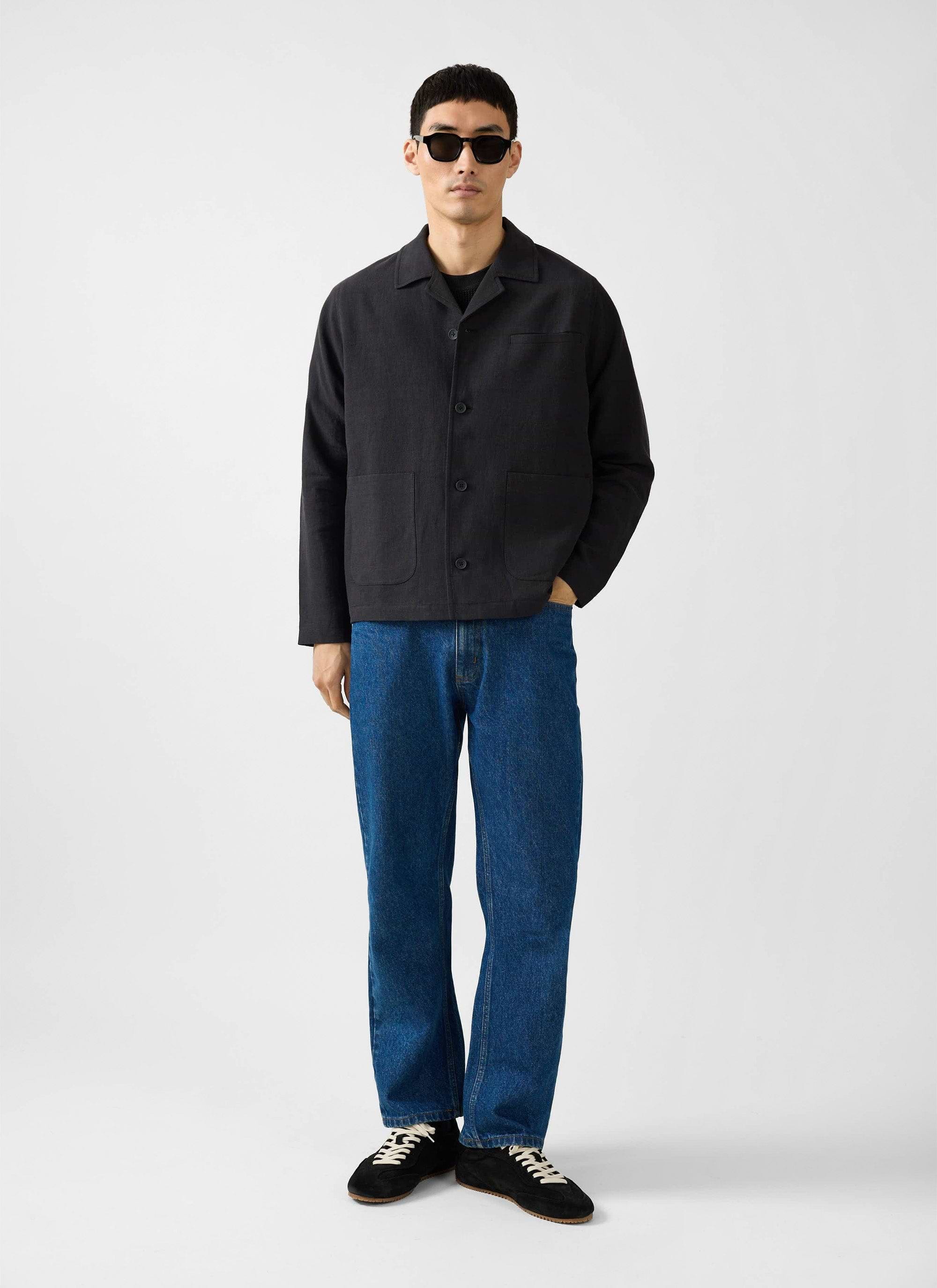 A full length shot of a model wearing the Percival black linen overshirt, blue jeans, sunglasses and black trainers.