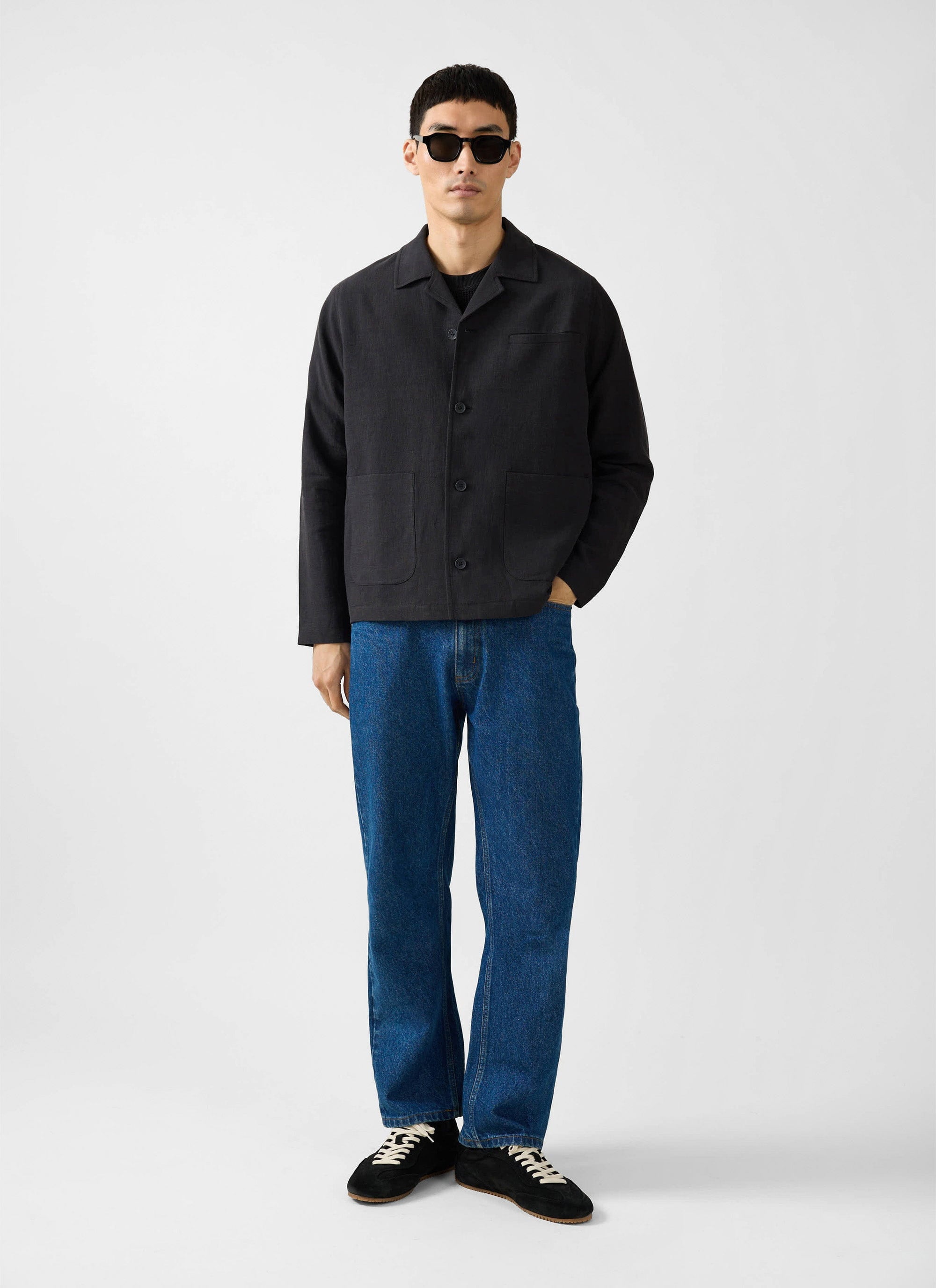 A full length shot of a model wearing the Percival black linen overshirt, blue jeans, sunglasses and black trainers.