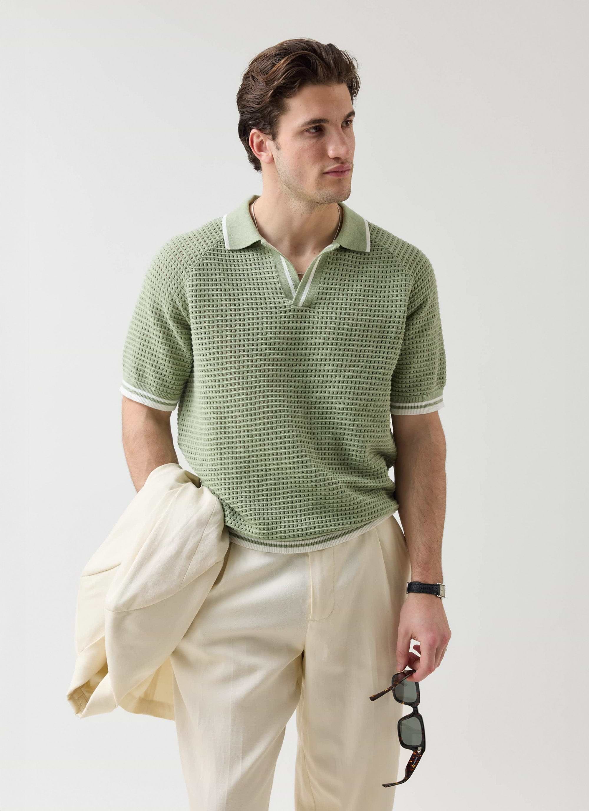 A man models the Percival Tennis Boxy Knitted Polo in sage with cream trousers, holding a jacket and sunglasses.