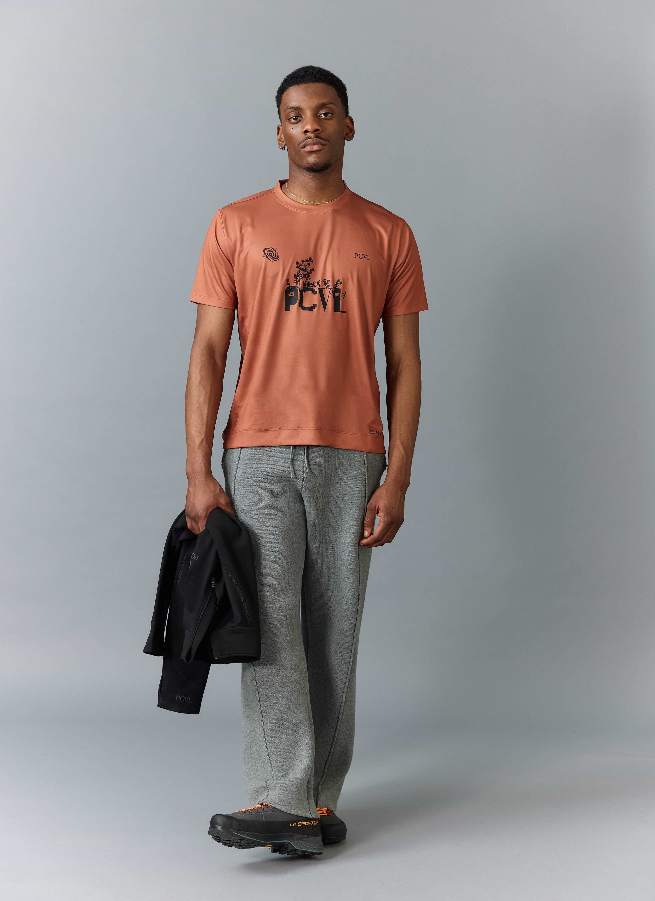 A model wears the Percival PCVL Printed Performance Top in rust, styled with grey trousers and holding a black jacket.