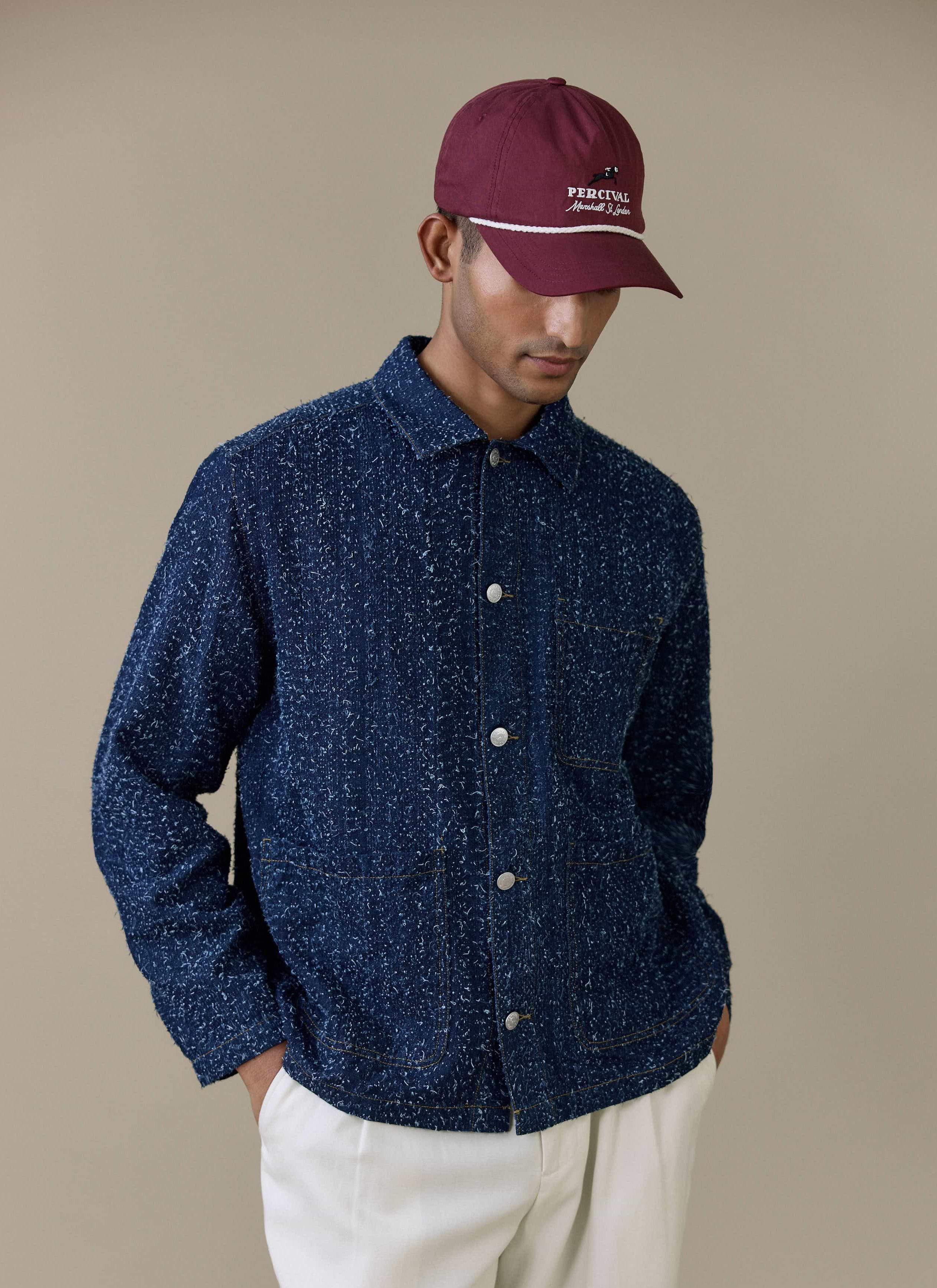 A model wearing the navy Parsons Chore Jacket and red Tournament Soft Trucker Cap from Percival.