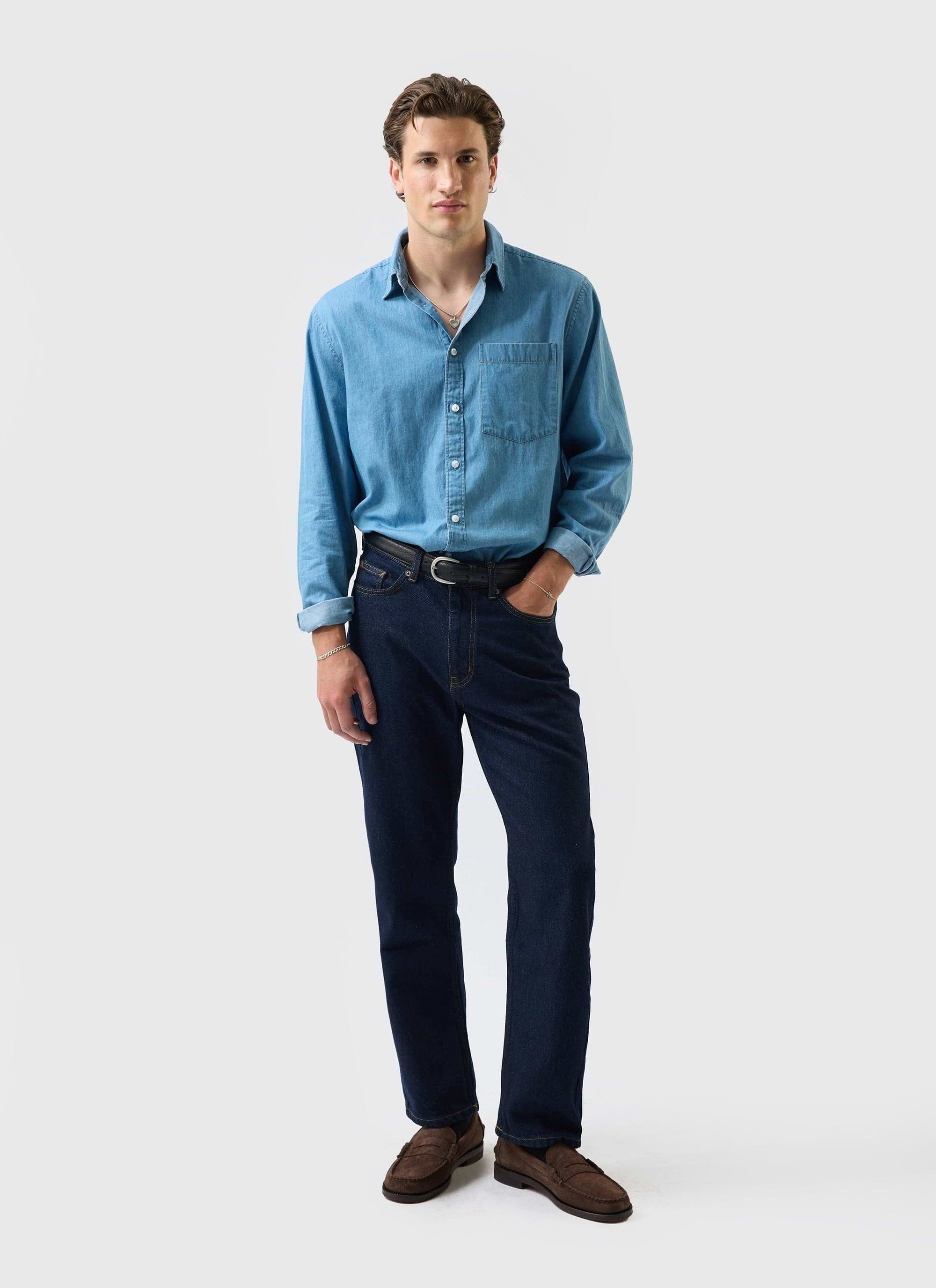 A man models the Percival Marco Pearce Shirt in light wash blue denim, paired with dark jeans and brown loafers.
