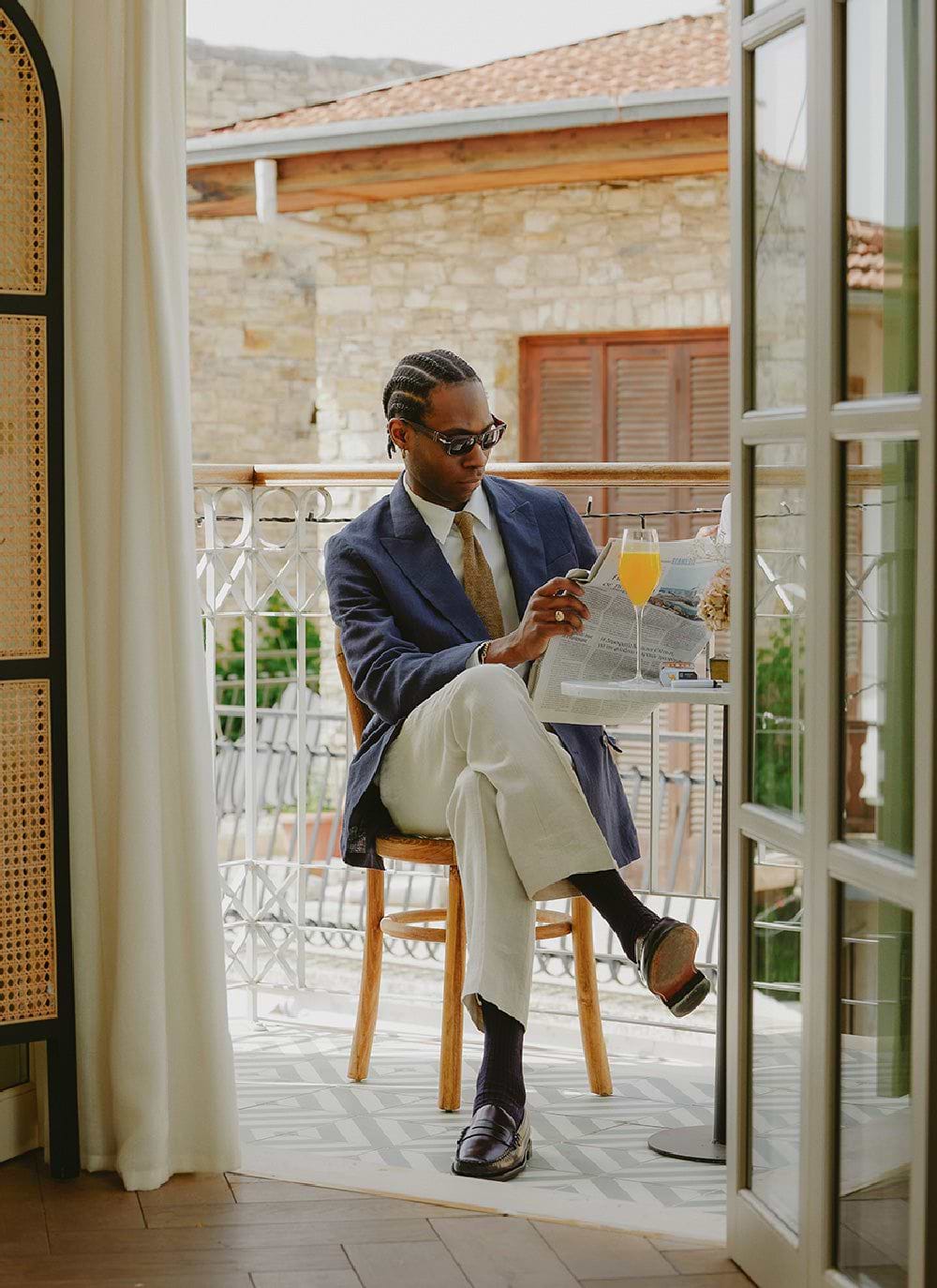 A man sits on a balcony reading a newspaper, wearing the Percival navy linen double-breasted blazer with cream trousers.