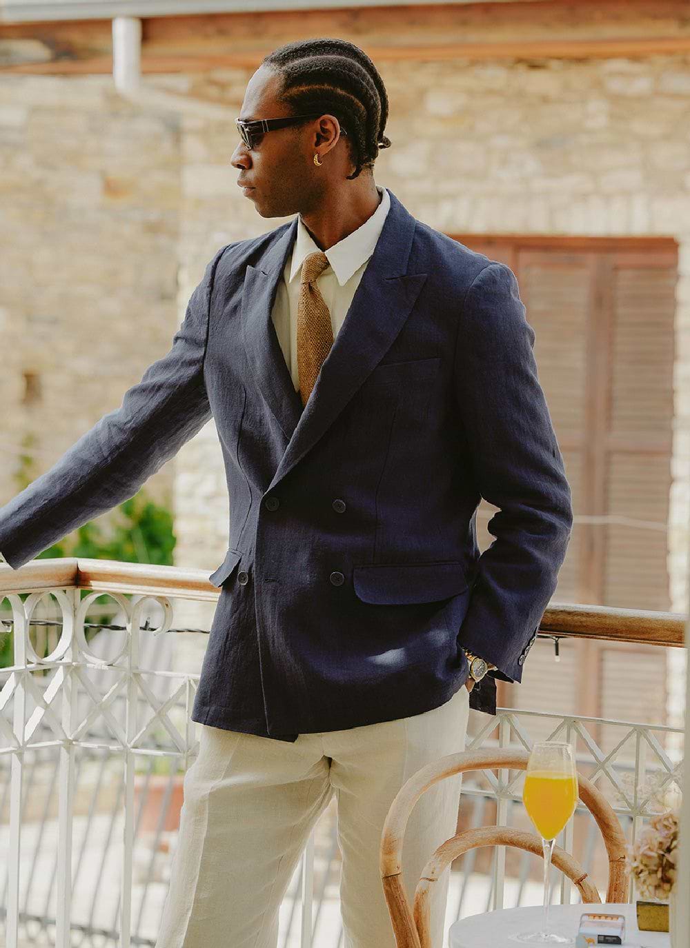 A man on a balcony wears the Percival navy linen tailored double breasted blazer with cream trousers and a brown tie.