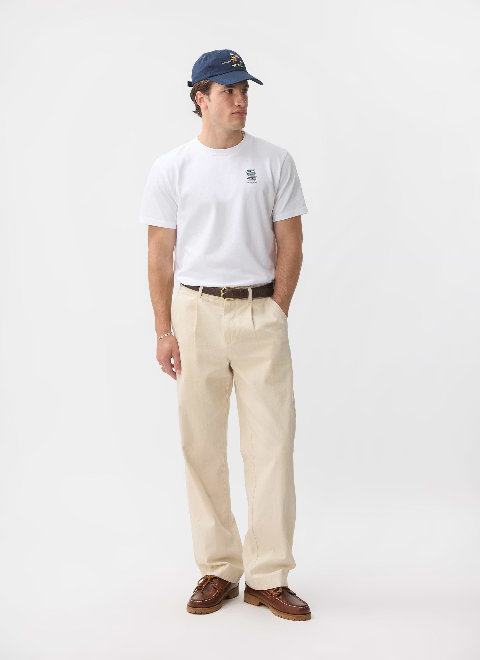 A man models the white Percival Duke T Shirt, styled with cream trousers, brown boat shoes, and a navy cap.