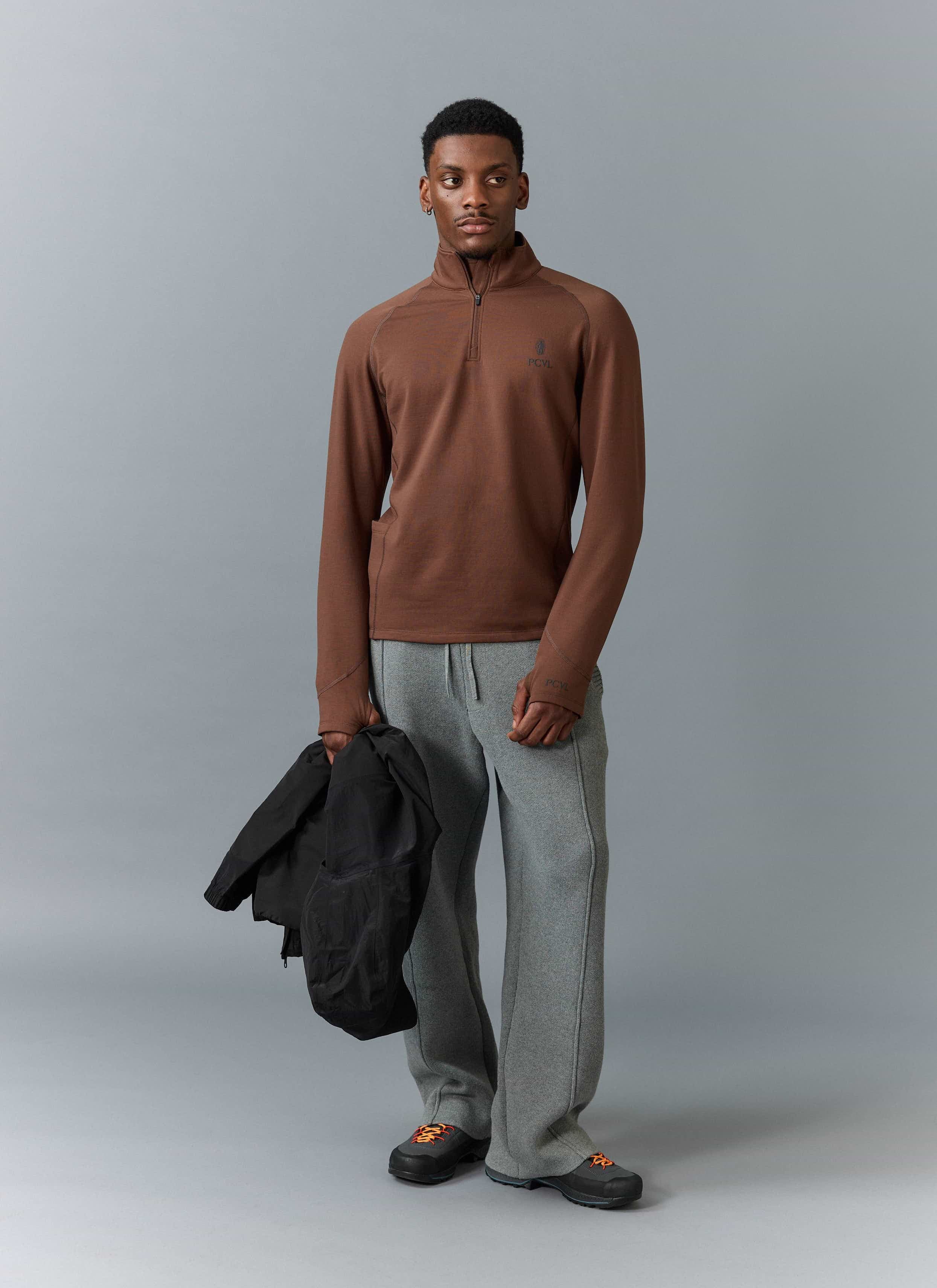 A model wears the Percival PCVL Base Layer Quarter Zip in rust, styled with grey trousers and holding a black jacket.
