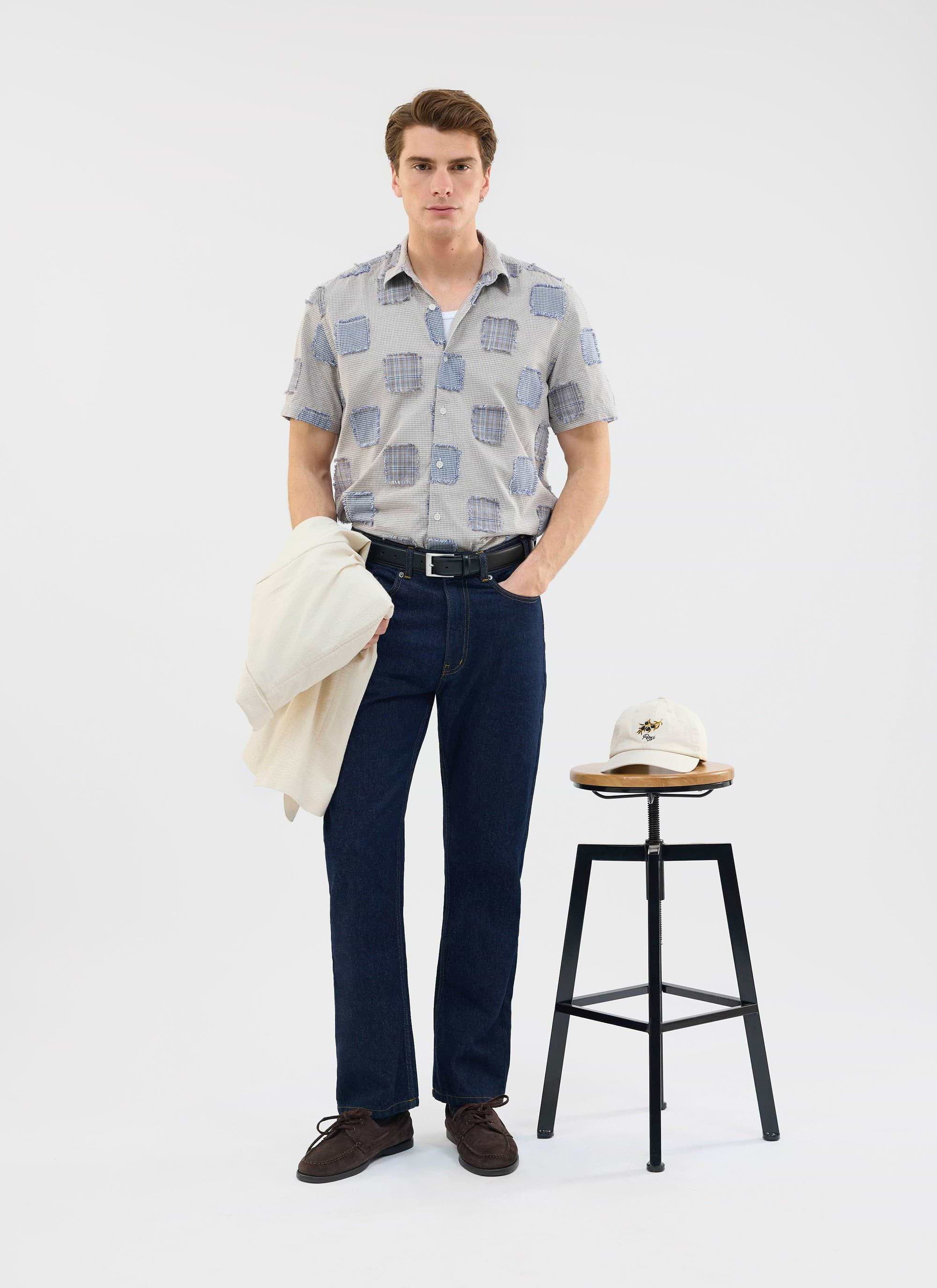 A model wears the Percival Bruno Boxy Clerk Shirt in grey, a patchwork design, paired with dark denim jeans.