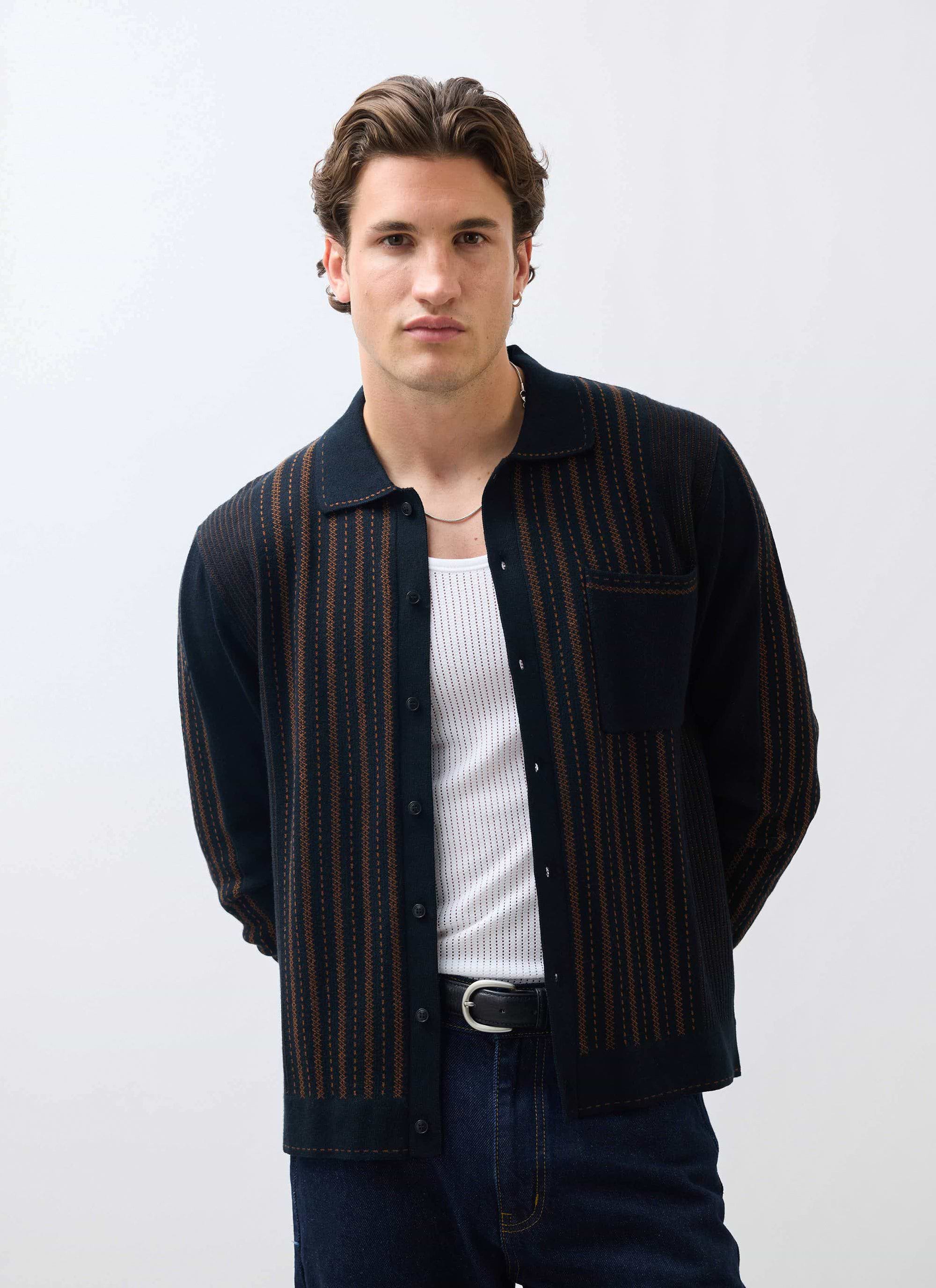 A model wears the Percival Appendix Knitted Shirt in black and brown, styled open over a white vest and jeans.